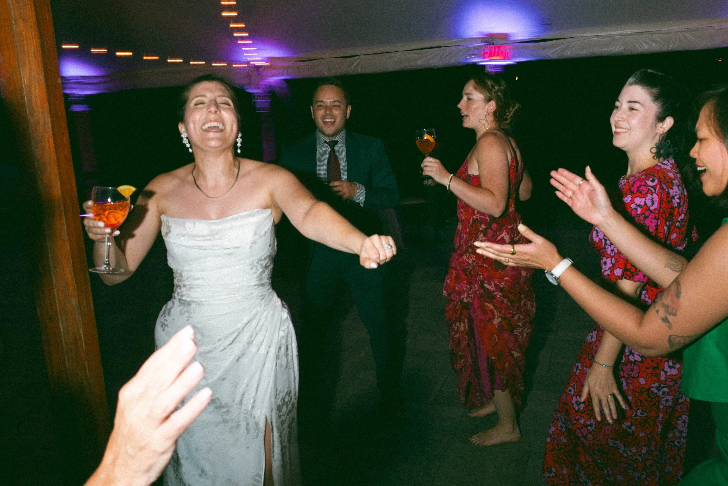 Bride smiiling and dancing with a red cocktail in her hand, surrounded by other guest dancing and drinking. 