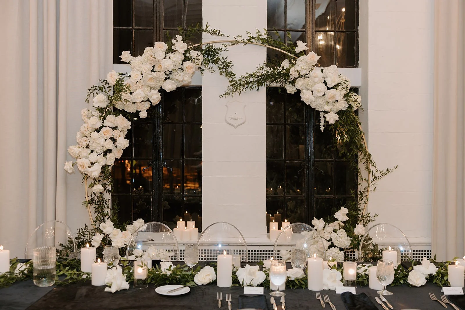 Elegant dining table decorated with white flowers, candles, and glassware, with a floral arch and black window behind.
