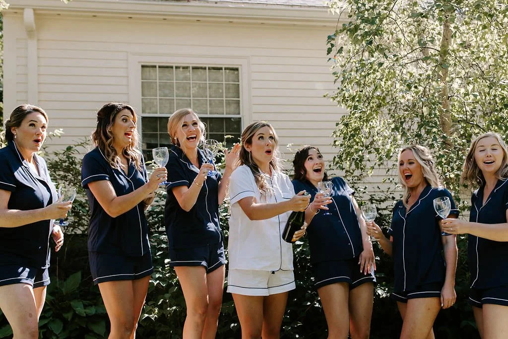 A group of seven women in navy pajamas celebrating outside, holding wine glasses, with a woman in white pajamas pouring champagne, in front of a house with trees and bushes.