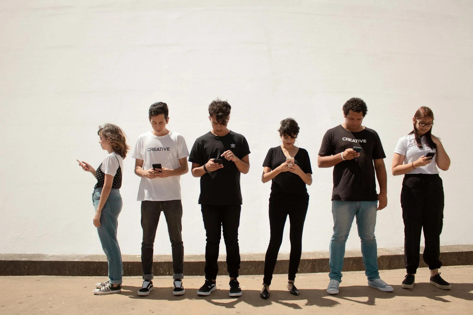 A row of young people standing against a wall, each looking at their own smartphone