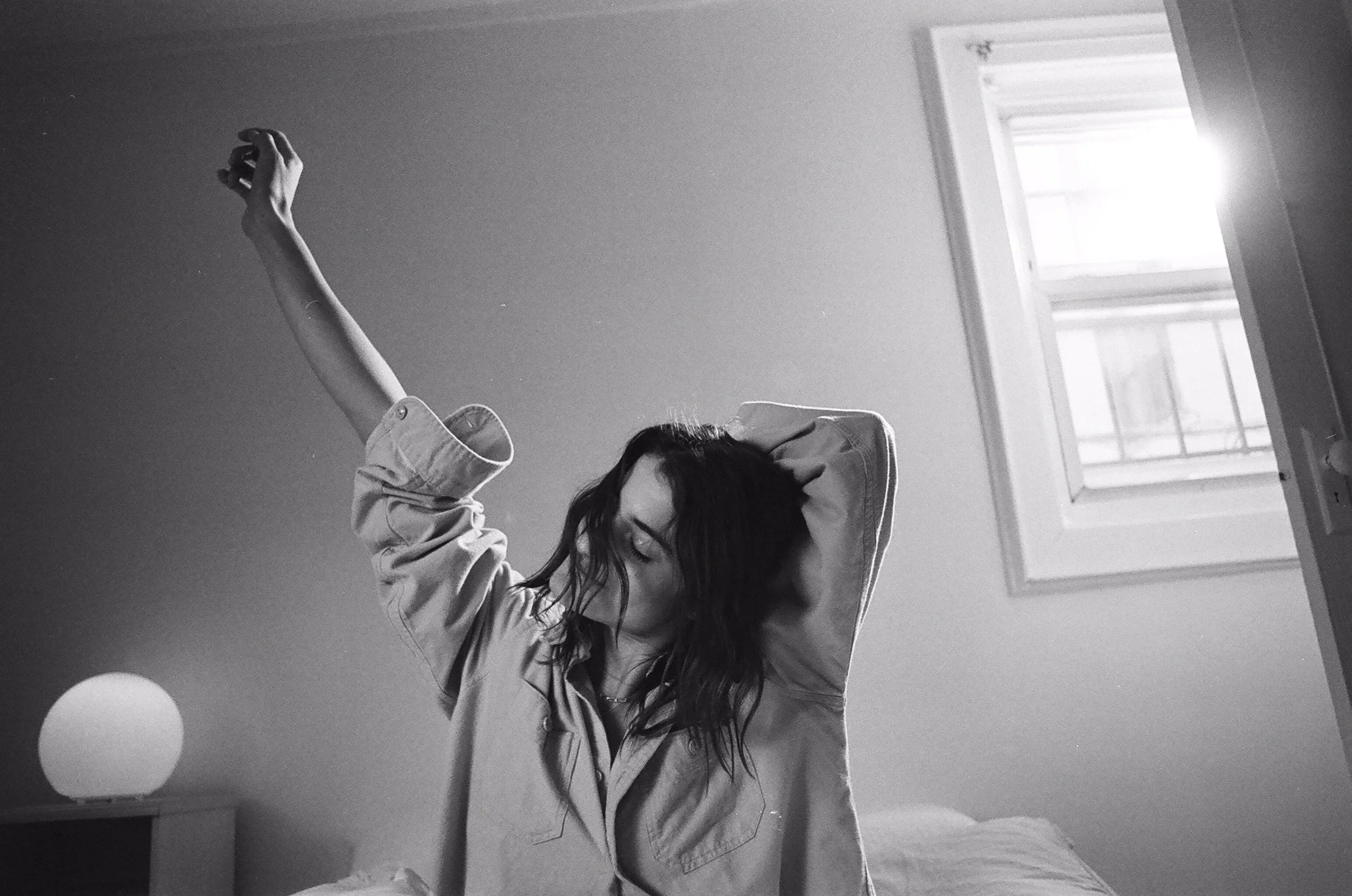 A woman with long hair stretching with one arm raised and the other behind her head in a bedroom with sunlight coming through a window.