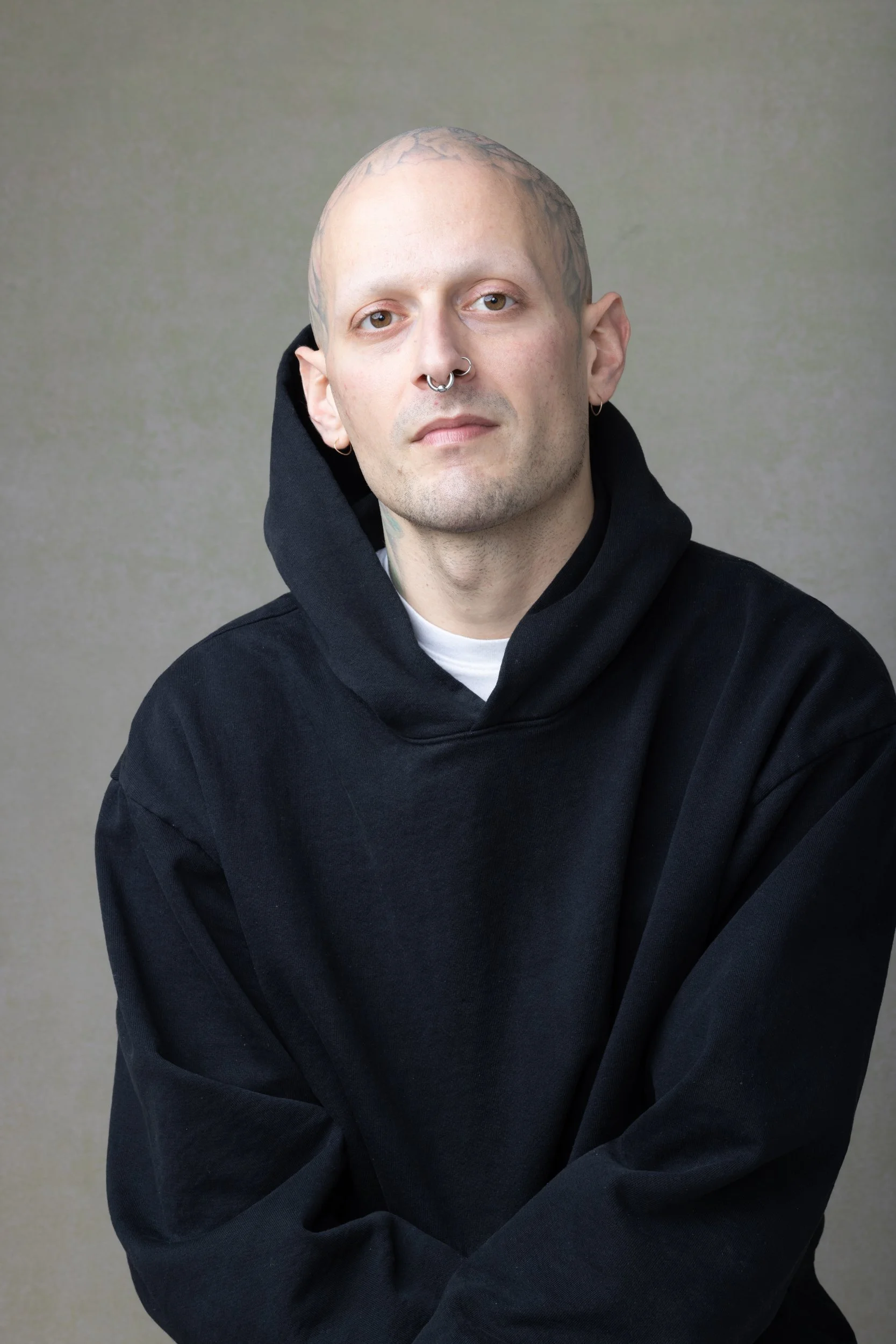 Portrait of a young man with a shaved head, multiple facial piercings, and tattoos on his scalp, wearing a black hoodie and standing against a neutral background.