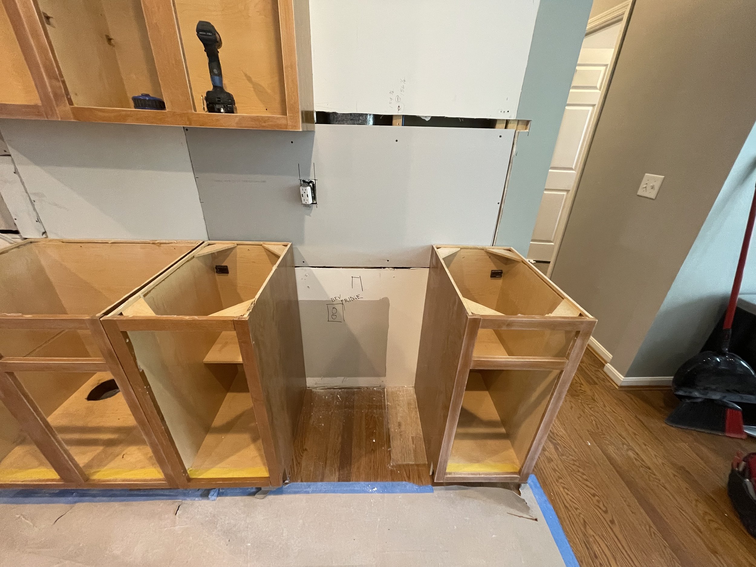 Kitchen renovation in progress showing unfinished wooden cabinets and a partial wall with drywall. Tools are on top of the cabinets, and the floor has protective blue tape and uncarpeted flooring. There are electrical outlets and markings on the drywall.