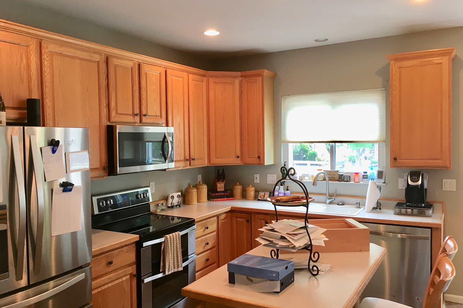 A kitchen with wooden cabinets, a stainless steel refrigerator, stainless steel microwave, black stove, and a small side fridge. There is a window over the sink, and various kitchen items on the counter including jars, paper, and a coffee maker. A kitchen island with papers and mail on top is in the center.