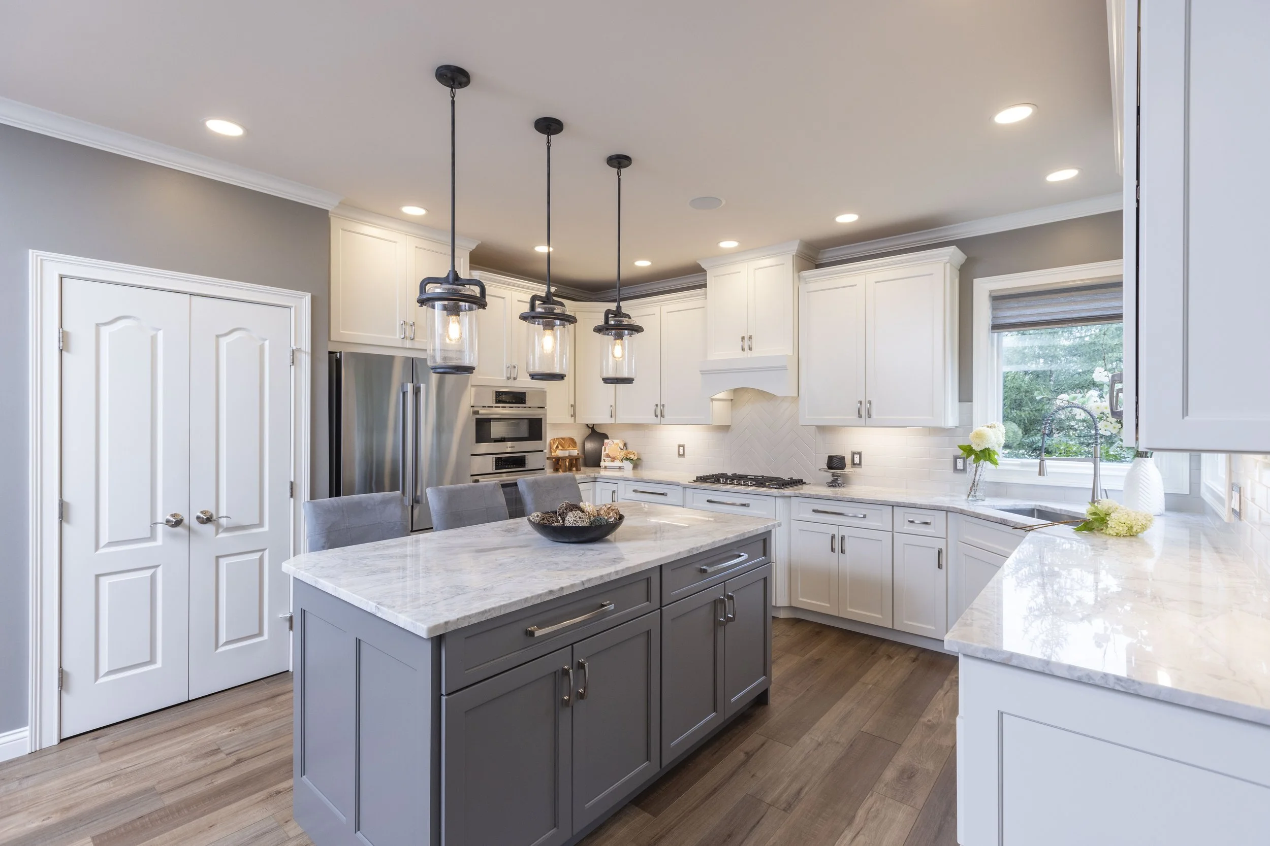 Modern kitchen with white cabinetry, a gray island, pendant lights, stainless steel appliances, and a window with a view of greenery.