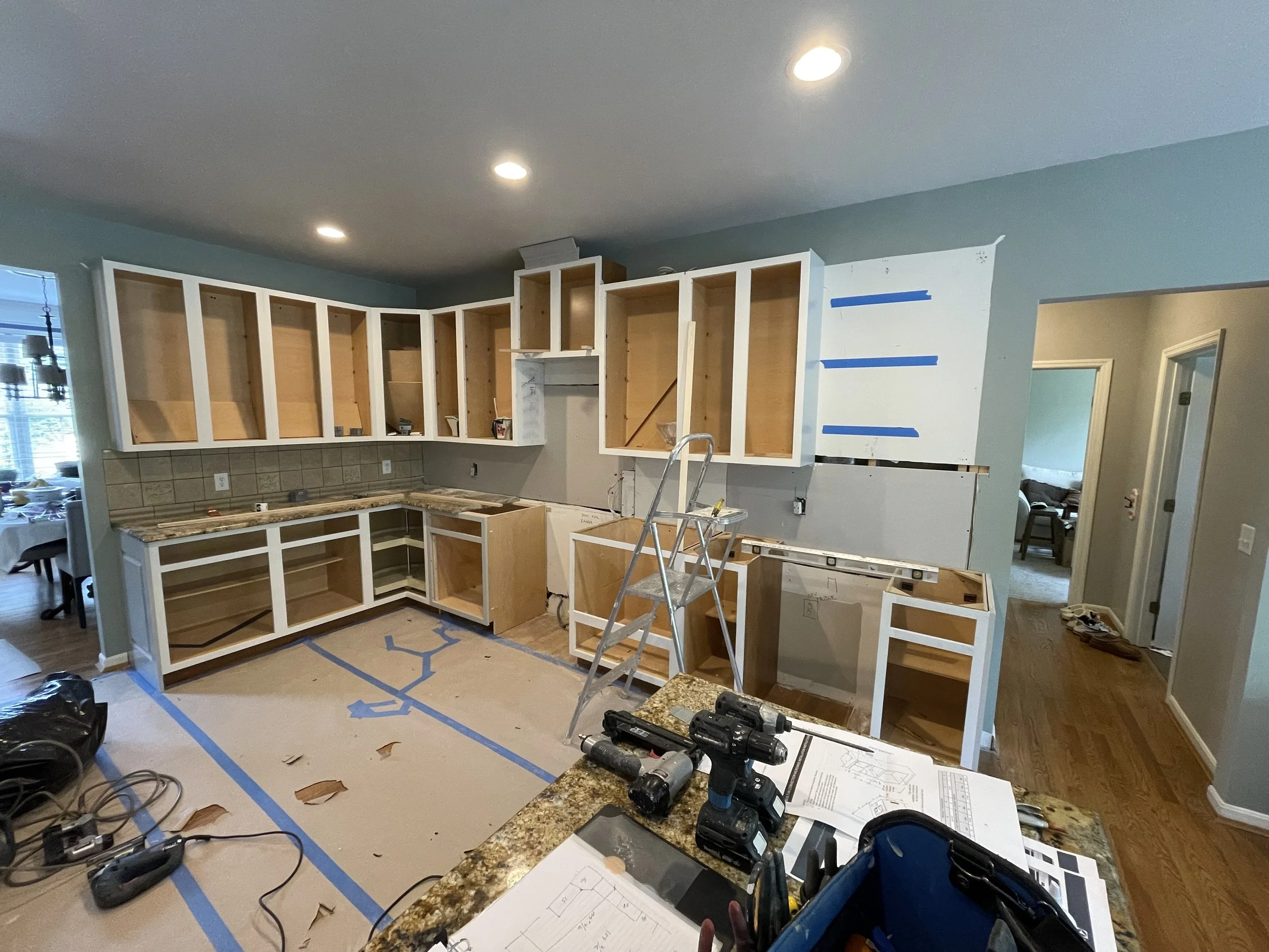 Kitchen mid-renovation with unfinished cabinets, construction tools, and blue painter's tape on the floor and wall.