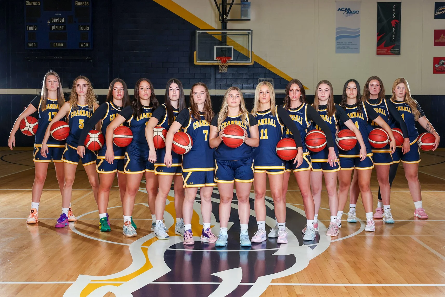 A girls' basketball team posing on an indoor basketball court, each player holding a basketball under their arm, wearing matching blue and yellow uniforms with 'Kendall' on the front.