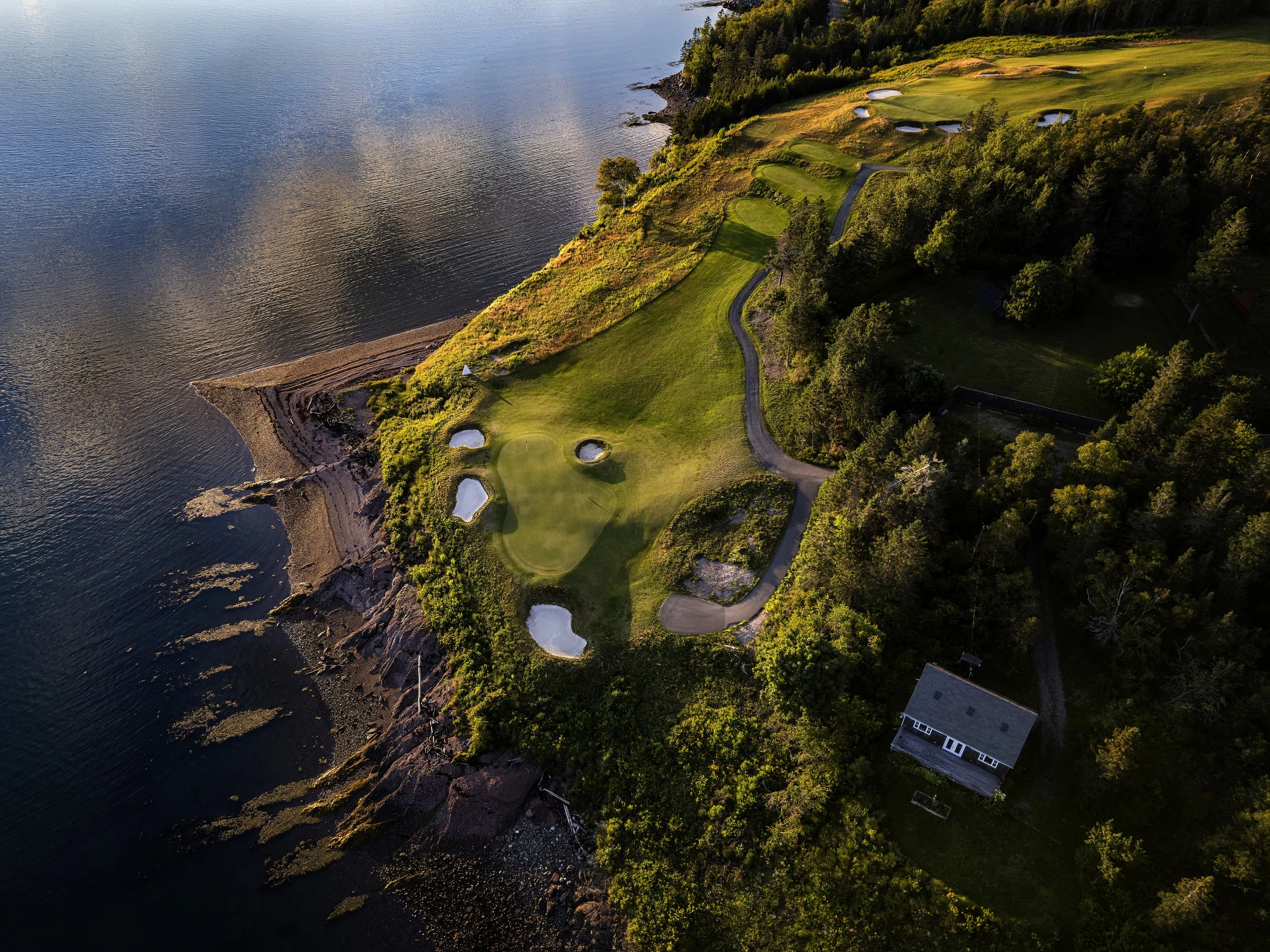 Aerial view of St Andrews golf course with green fairways, sand bunkers, and trees along the shoreline, with the ocean on the left.