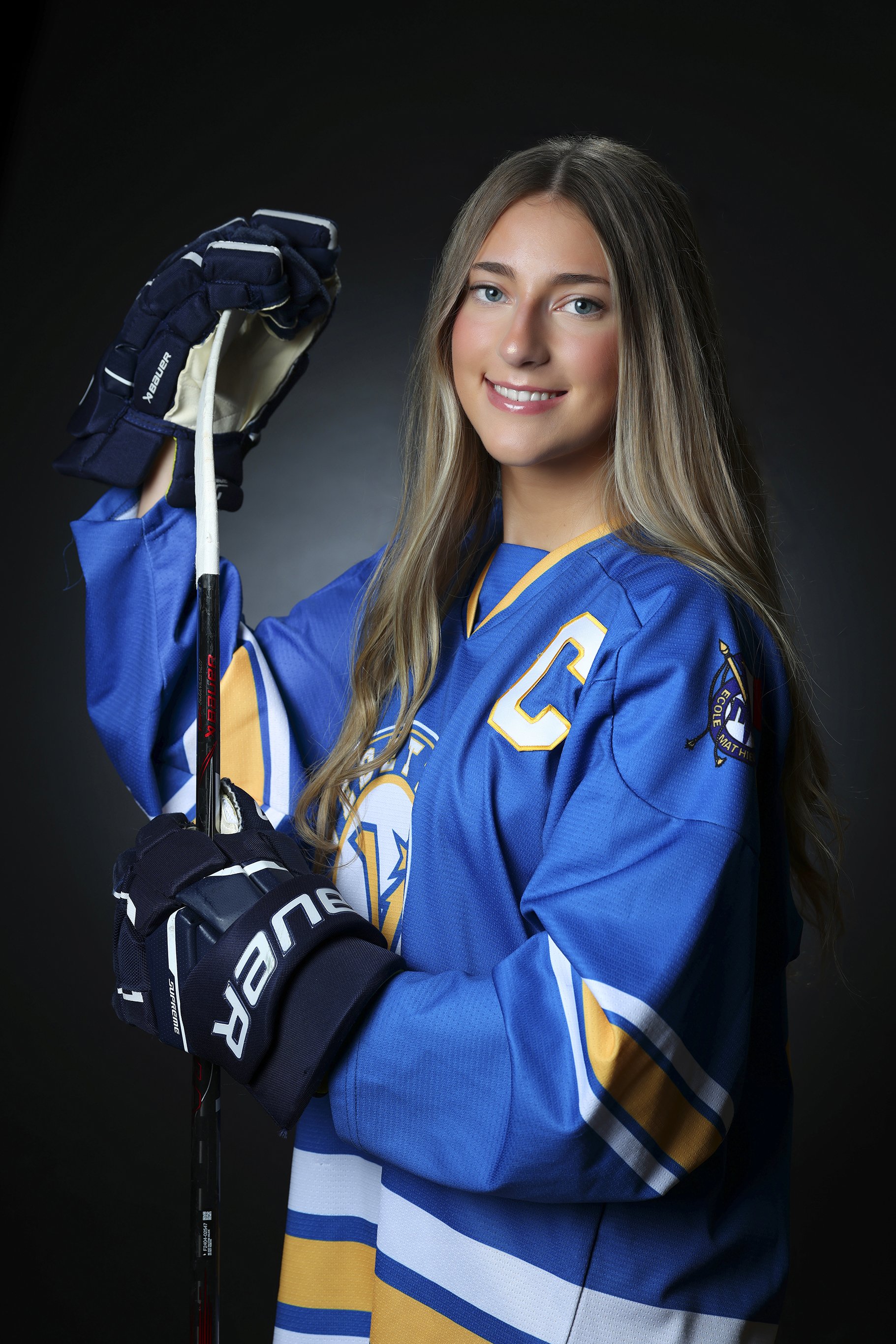 Young woman in hockey gear holding a hockey stick, smiling, wearing a blue and yellow hockey jersey with a 'C' on the front and a patch on the sleeve, against a dark background.