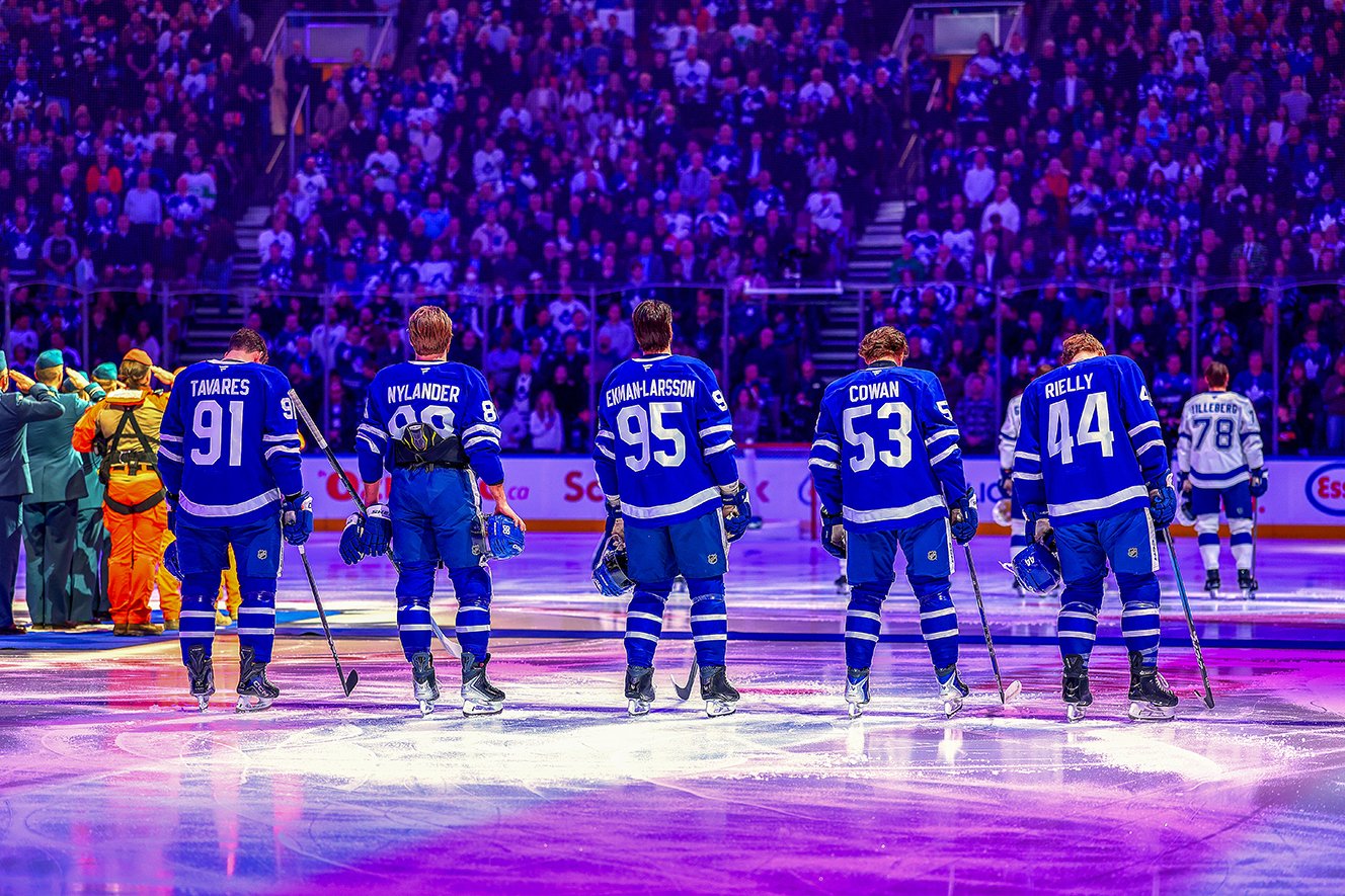 Hockey players in blue uniforms standing on ice rink before a game, with a crowd in the stands.