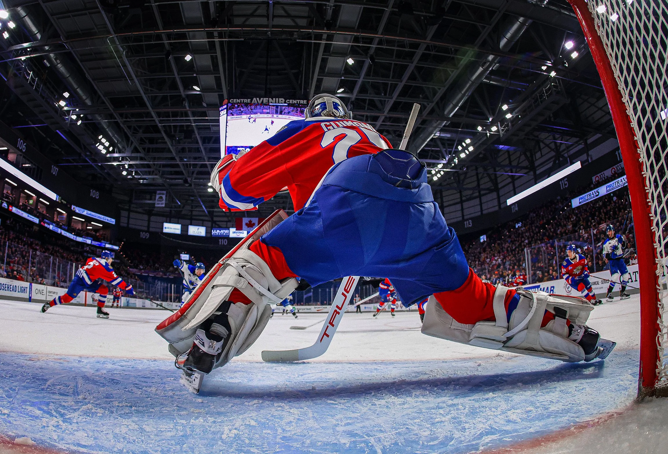 An ice hockey game seen from behind the goalie in front of the net, with players on the ice and a scoreboard above in an indoor arena.