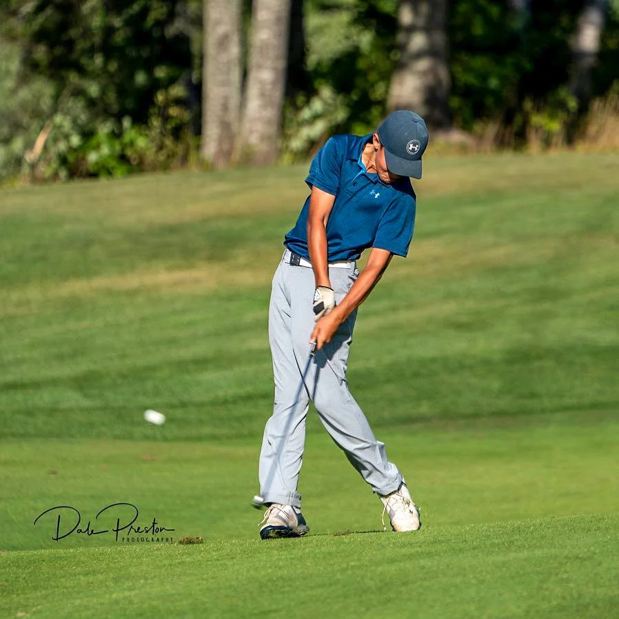 A young boy playing golf on a golf course, wearing a blue polo shirt, gray pants, a baseball cap, and golf gloves, preparing to putt the ball.
