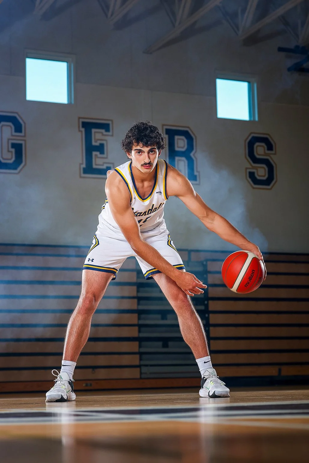 A young man in a basketball uniform crouches and prepares to dribble a basketball inside a gymnasium with the word 'GERS' on the wall behind him.