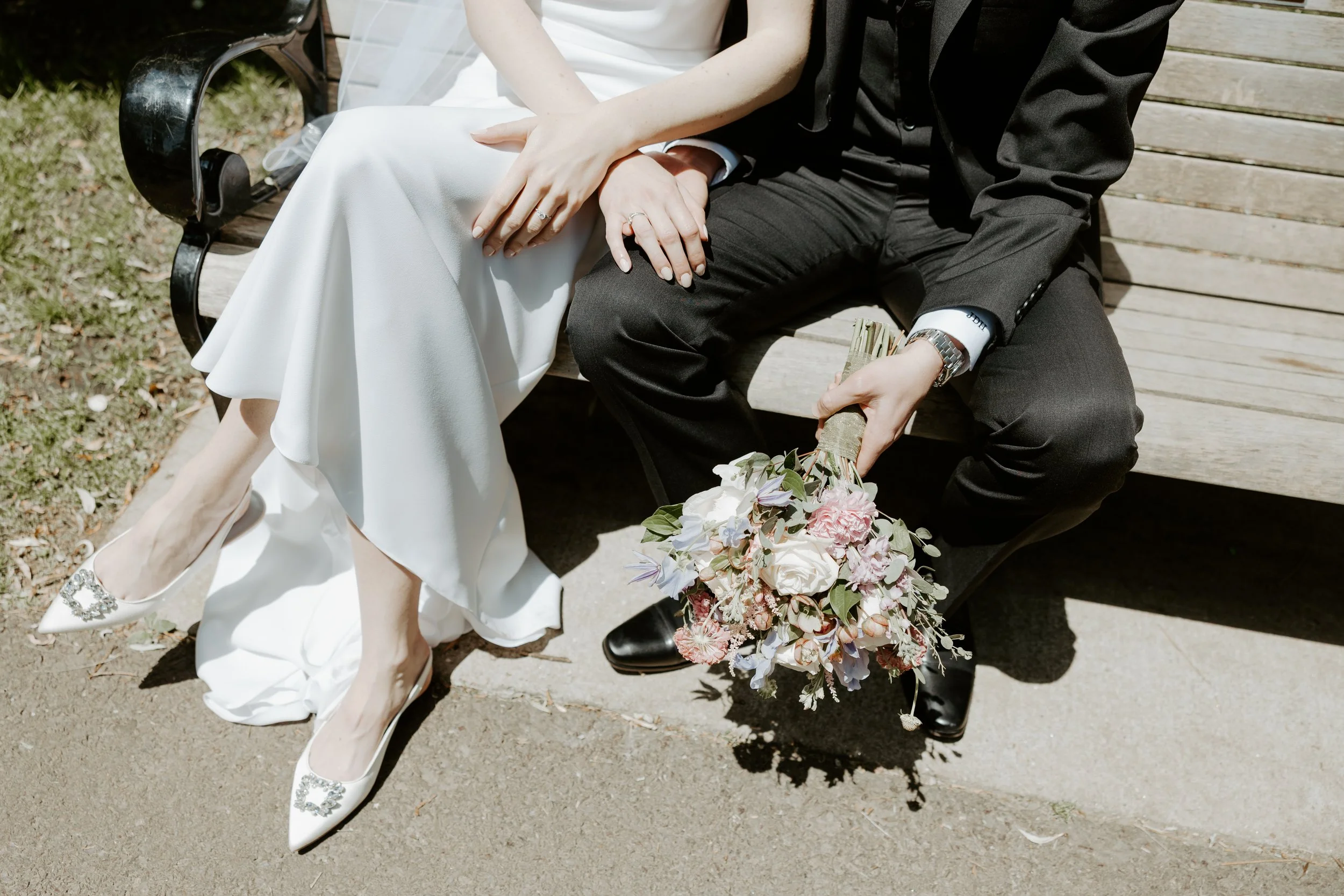 A couple sitting on a bench before heading to their elopement location, showing their shoes and a bridal bouquet