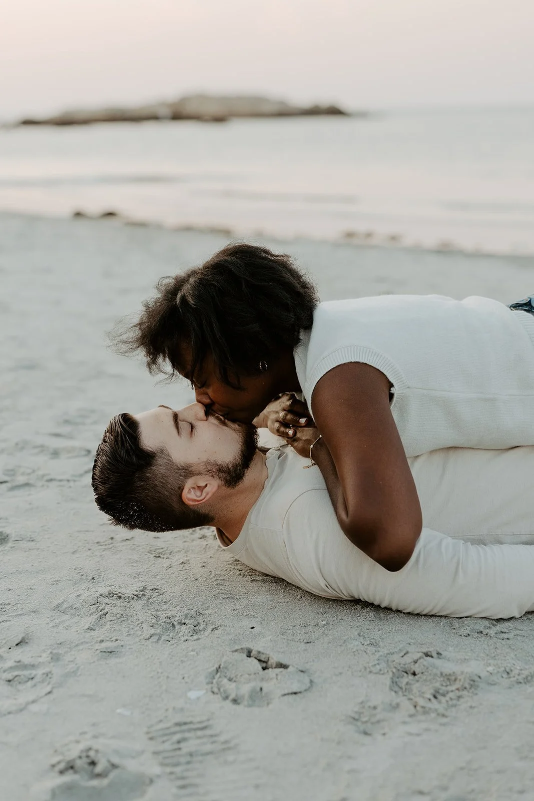 A couple kissing laying on the sand in their beach engagement photoshoot