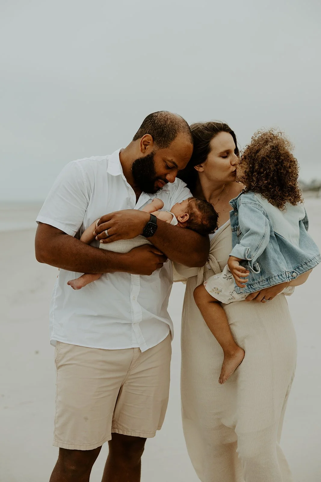 A mom kissing her daughter and dad looking at son during a beach family photoshoot
