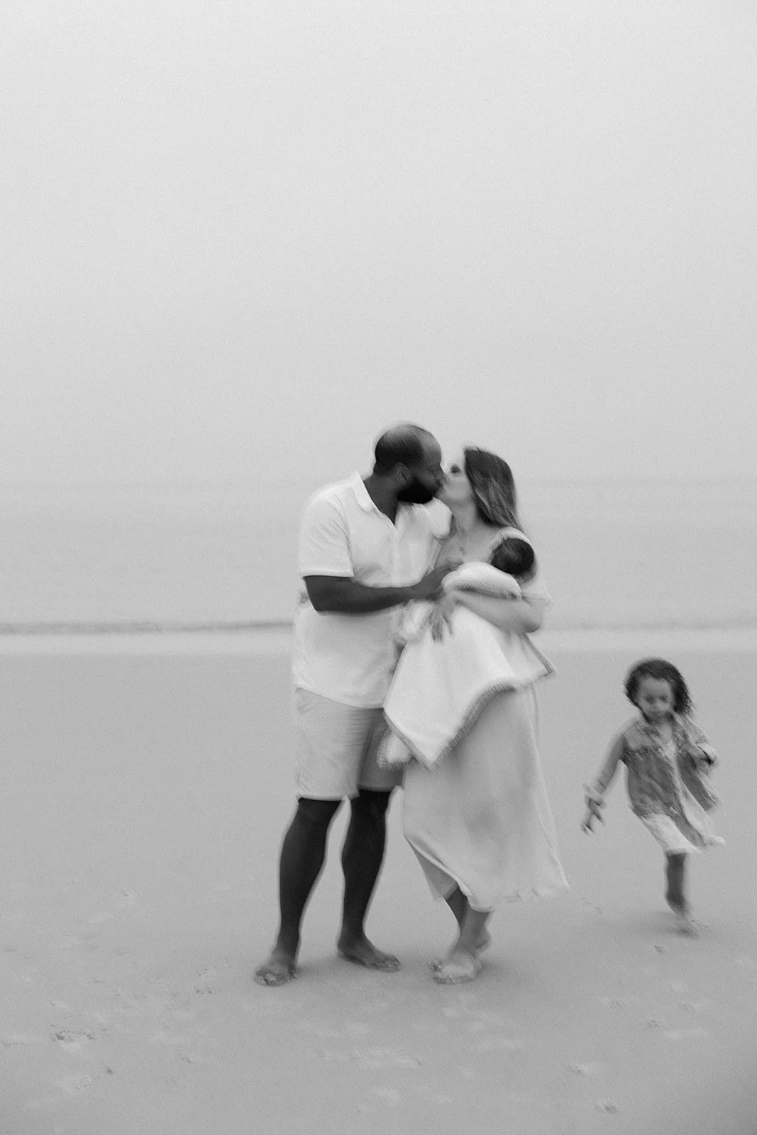 A couple kissing during a beach family photoshoot holding a baby while their daughter runs around them