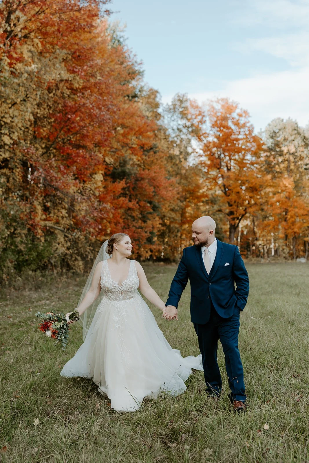A bride and groom walking through a field in front of fall leaves