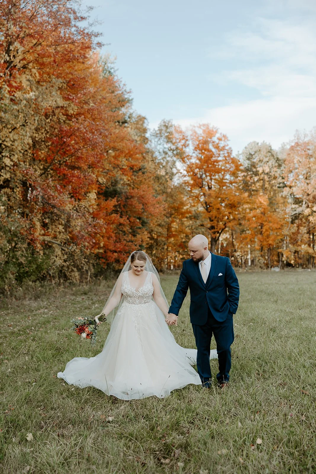 A bride and groom walking through a field in front of fall leaves