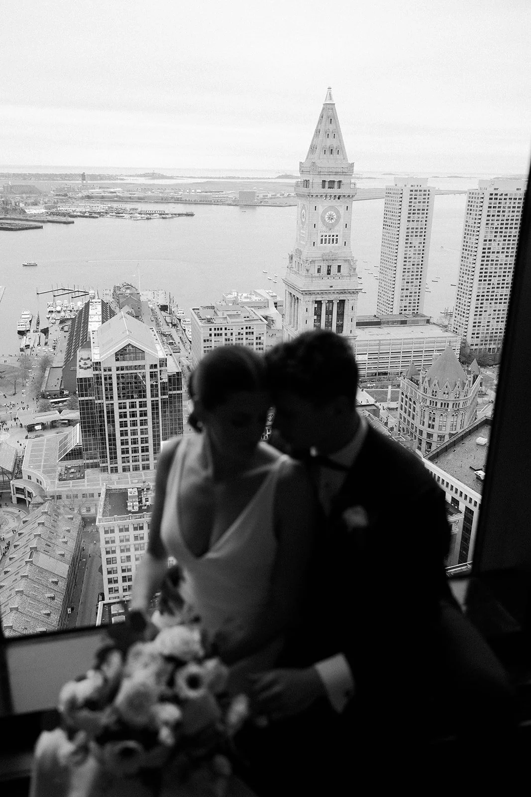 A black and white photo of a bride and groom looking out of the window from their wedding venue in Boston