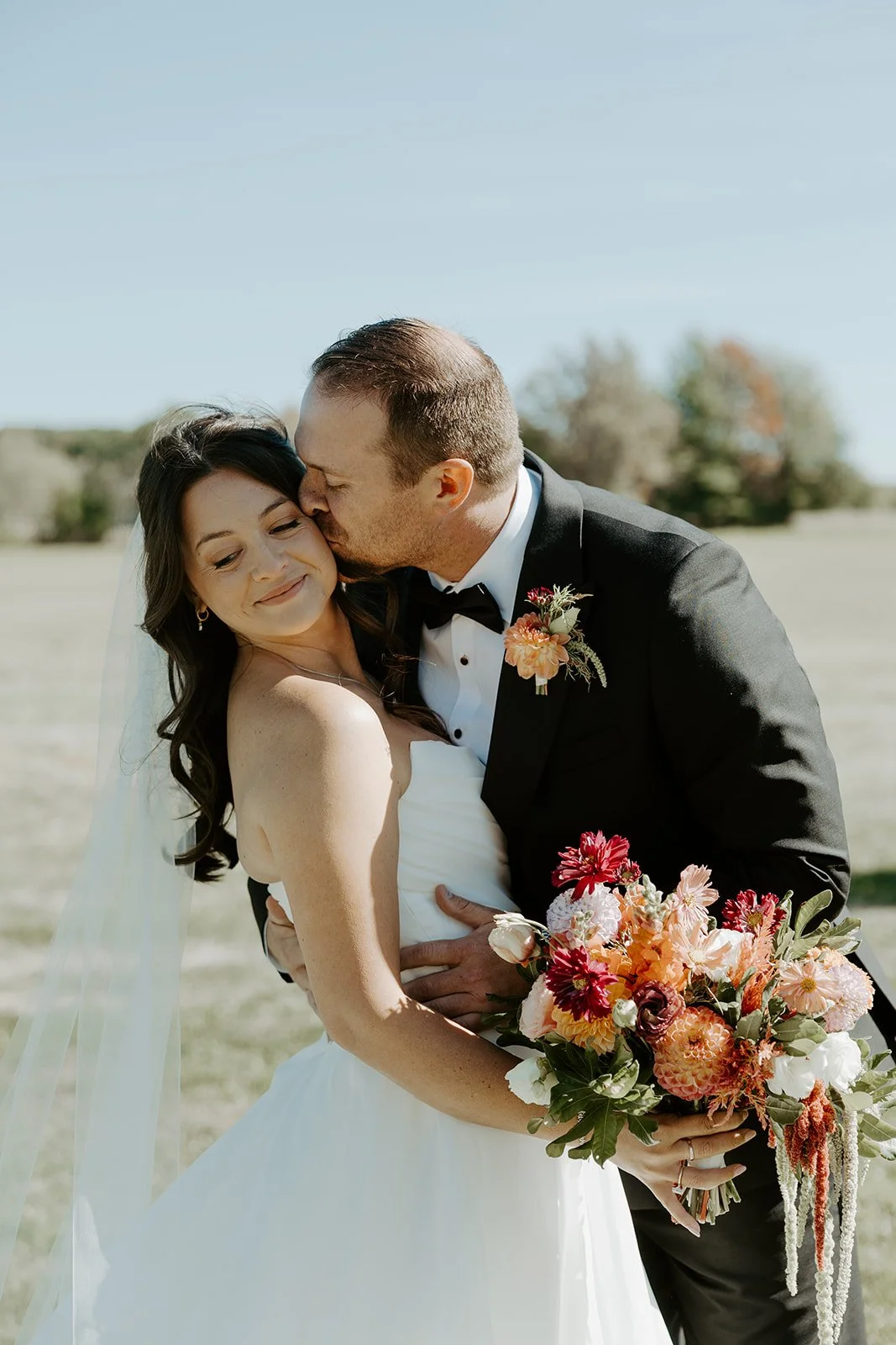 A groom kissing a bride on the cheek at a backyard wedding