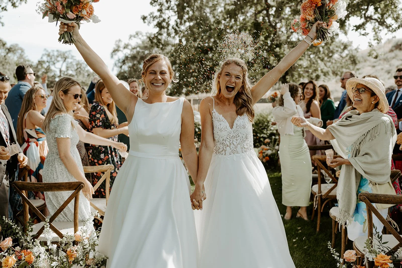 Two brides cheering at their whimsical wedding while wedding guests are throwing rainbow sprinkles