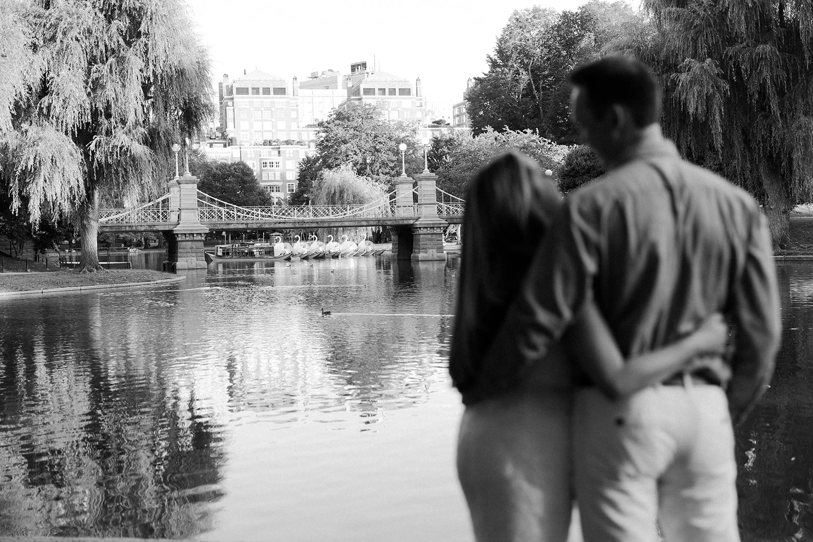 A black and white image of a couple standing in front of the Boston skyline from Public Garden
