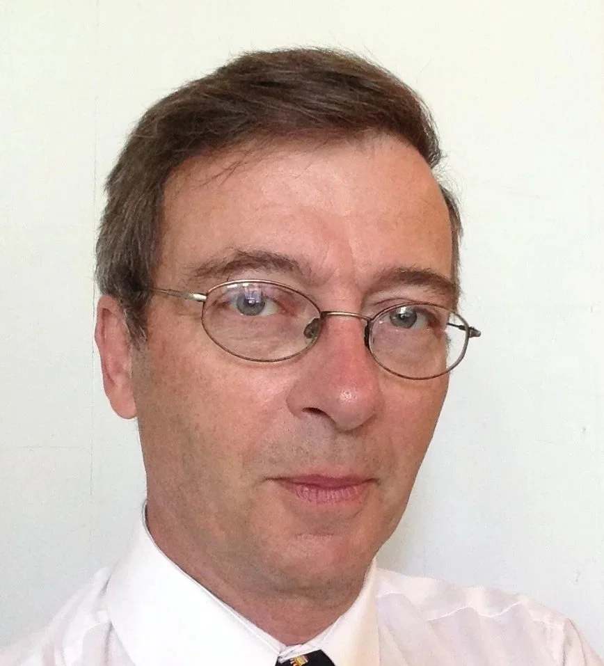 A man with glasses and light brown hair, wearing a white collared shirt and a striped tie, taking a close-up selfie against a plain white wall.
