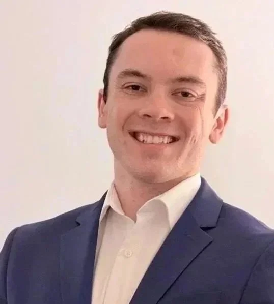 Portrait of a young man smiling, wearing a dark blue suit and white shirt, against a plain light background.
