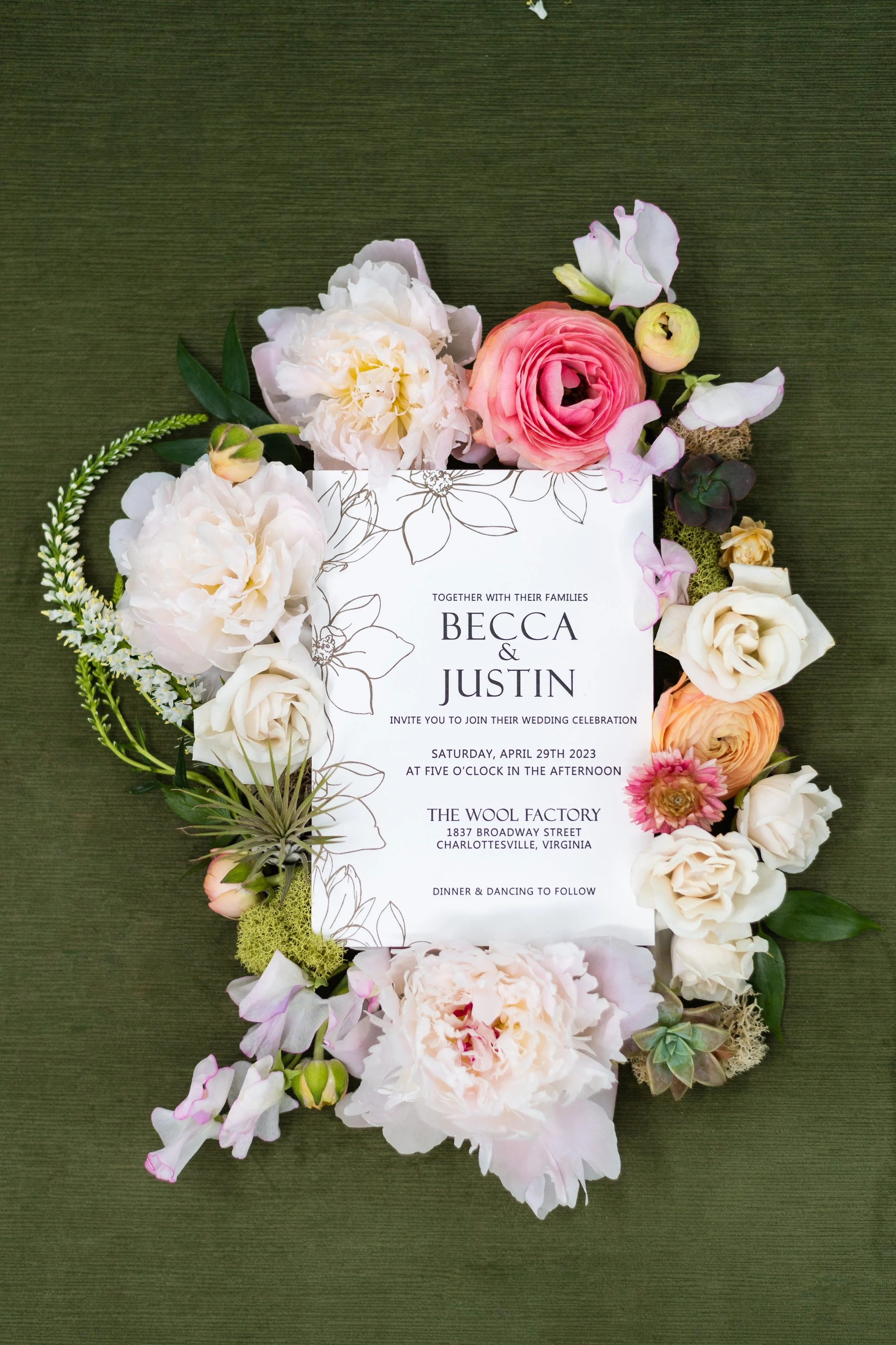 Wedding invitation surrounded by pink, white, and peach flowers with green foliage, on a green fabric background.