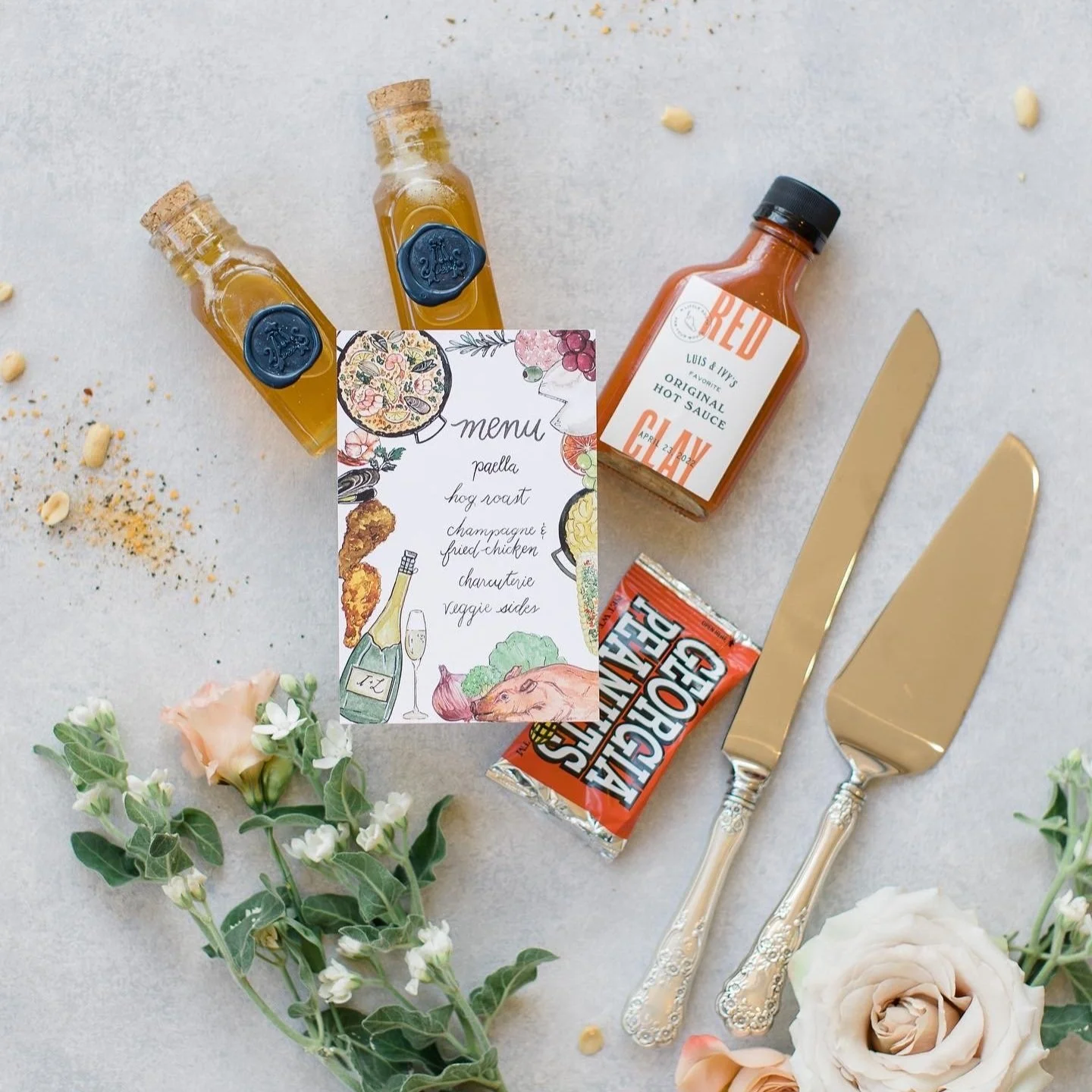 A flat lay of a gourmet meal setting featuring a handwritten menu, two small bottles of golden sauce, a bottle of hot sauce, a chocolate bar, a gold-handled knife and fork, and decorative flowers and greenery on a white surface.