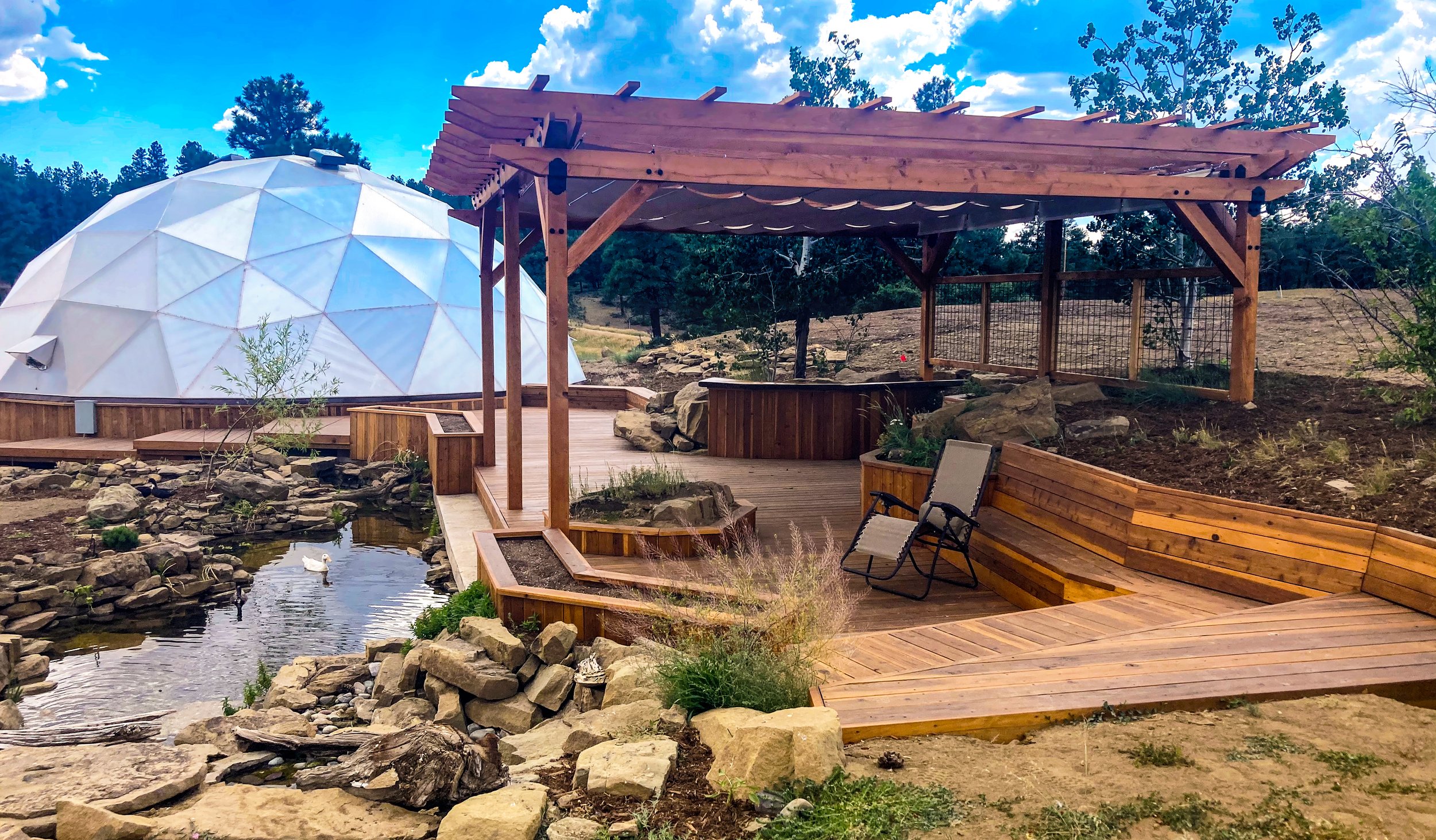 A backyard scene with a geodesic dome, a wooden deck, a hot tub, a small pond with ducks, and a wooden pergola structure under a partly cloudy sky.