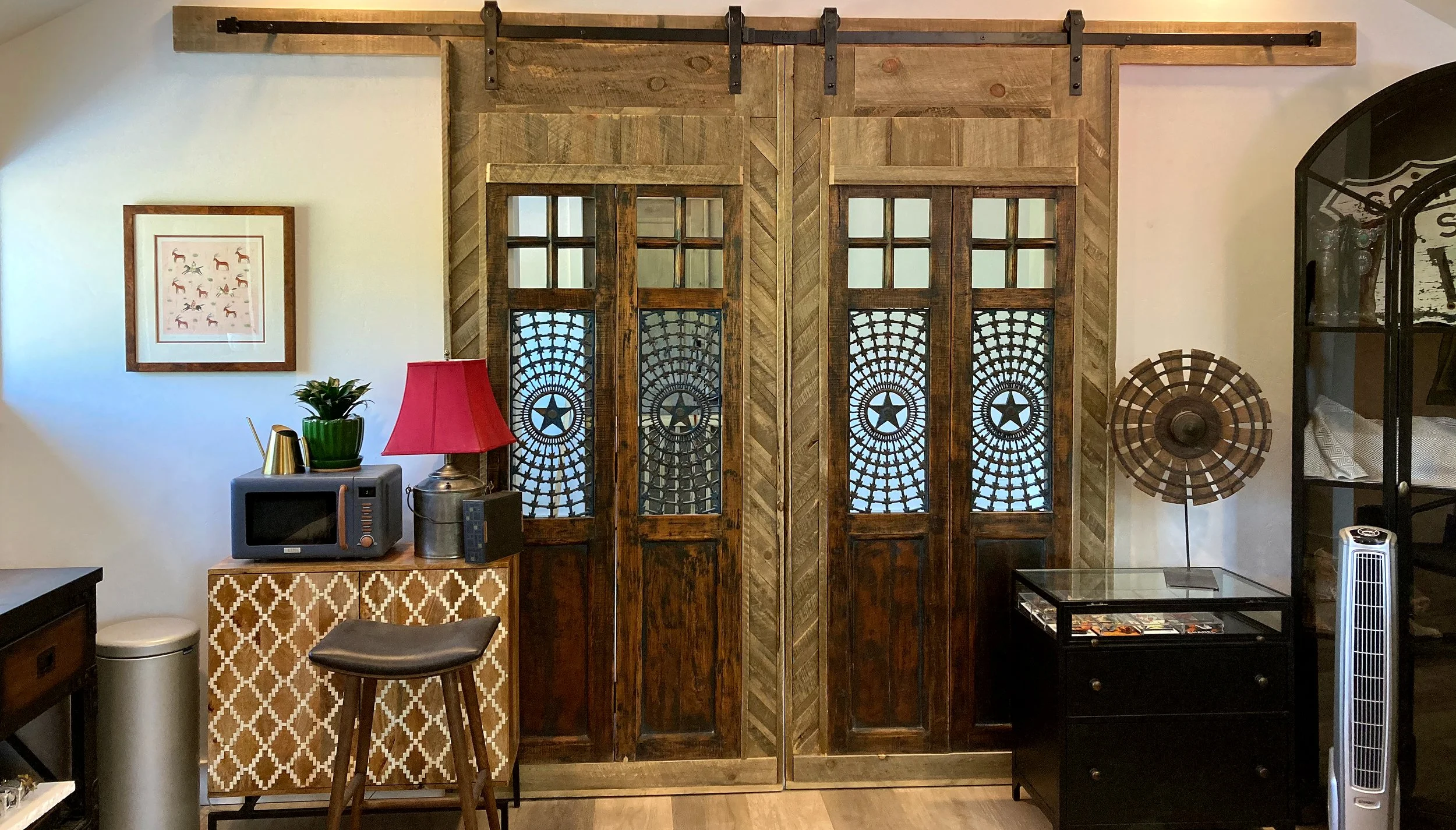 Decorative interior with wooden sliding barn doors featuring star and web design, small furniture, a framed art piece with horses, a potted plant, a pink lampshade, microwave, a gold watering can, lamp, remote controls, a lounge chair, a black cabine