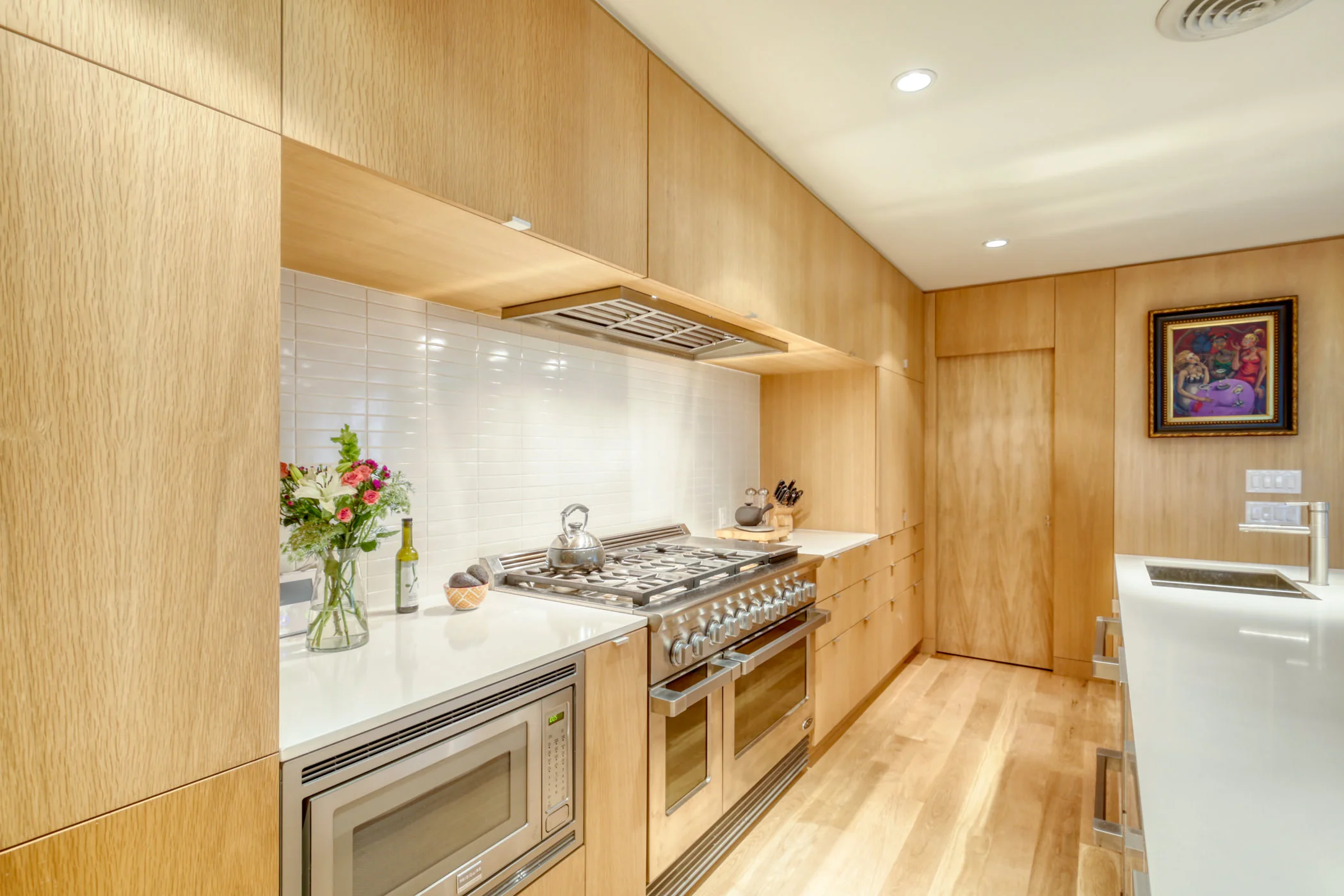 A modern kitchen with wooden cabinets, a stove, a microwave, a white countertop, a kitchen sink, flowers, a bottle, and a colorful framed painting on the wall.