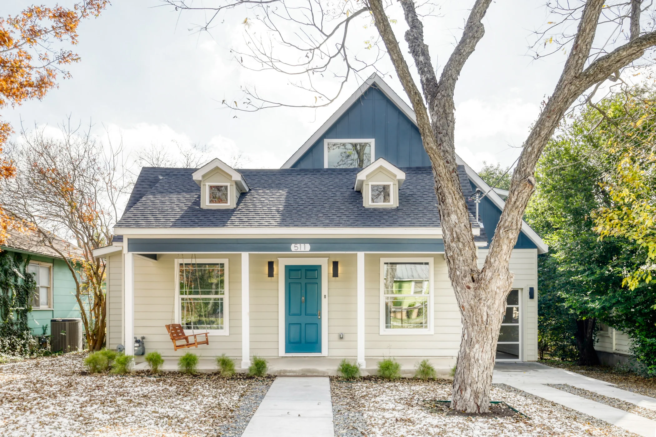 Front view of a modern two-story house with a blue front door, white siding, and dormer windows in a residential neighborhood. The yard has a concrete pathway, small bushes, a porch swing, and a large tree. Fall foliage is visible on the left.