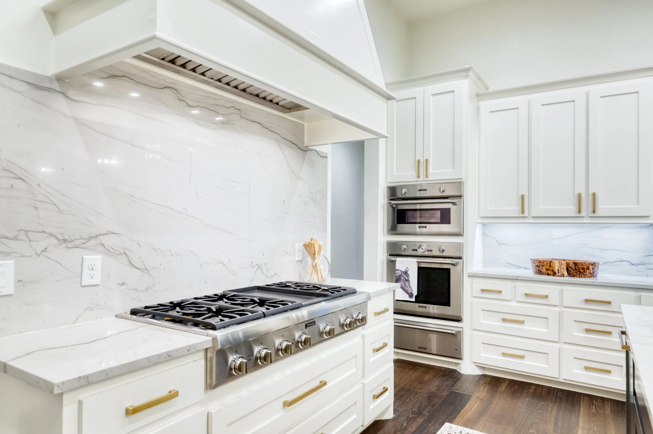 Kitchen with white cabinets, stainless steel oven and microwave, marble backsplash and countertops, dark hardwood floor, and a small wooden bowl on the counter.