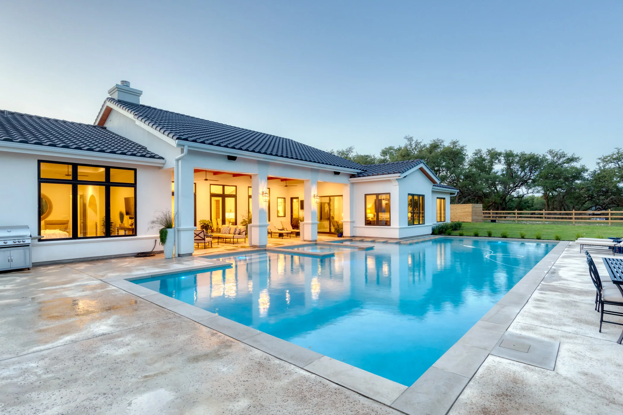 A backyard with a large swimming pool outside a white house with large windows and a covered patio area, surrounded by green grass and trees.
