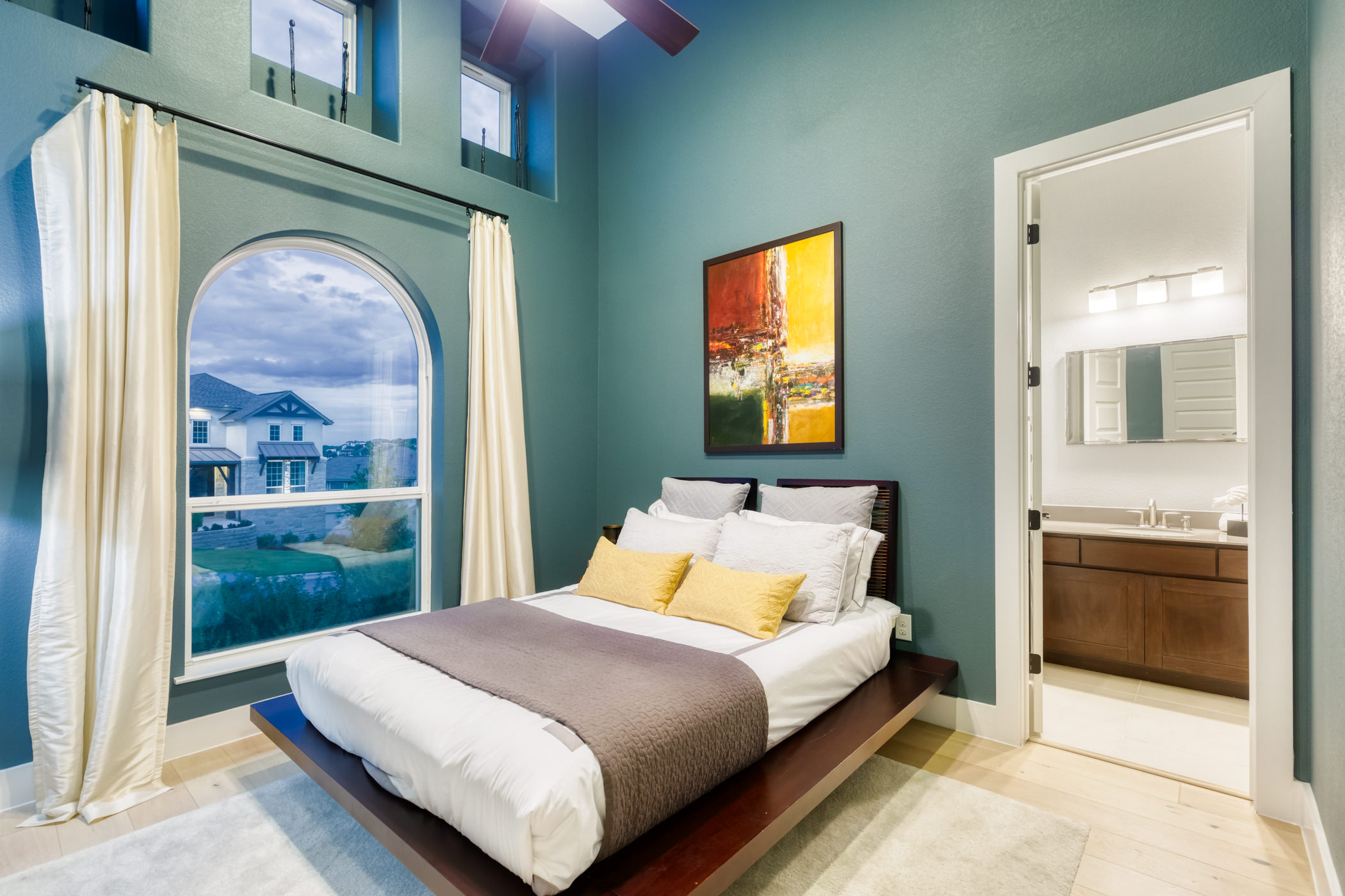 A modern bedroom with a large window, bed with multiple pillows and a blanket, an abstract painting on the wall, and an en suite bathroom visible through an open door.
