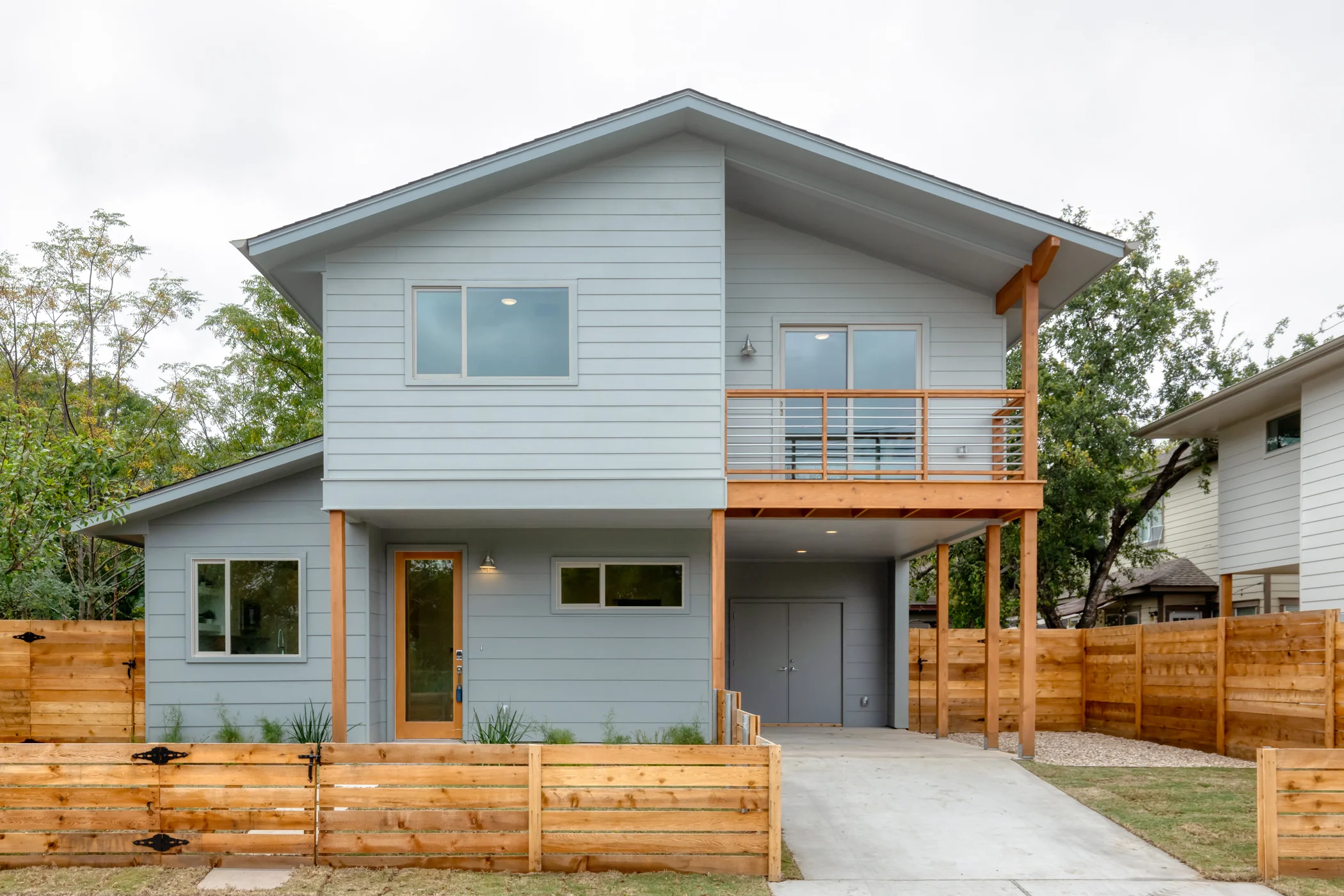 Modern two-story house with light blue exterior, wooden balcony and fencing, and a driveway in front.
