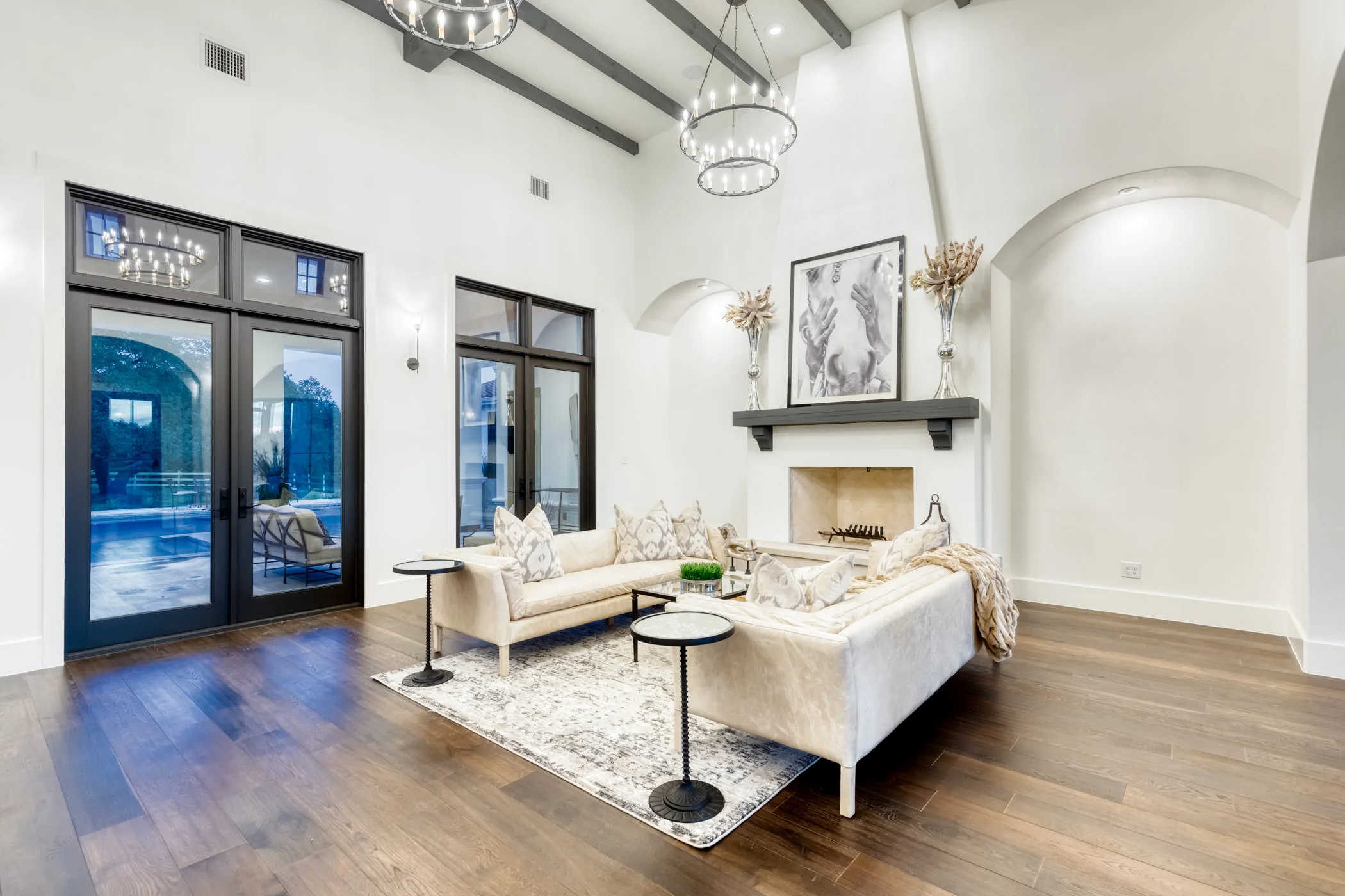 Living room with white walls, dark wood floors, two beige sofas, black side tables, a fireplace with a black mantel, modern black chandeliers, decorative vases with dried flowers, black framed windows, and a black and white art piece above the firepl