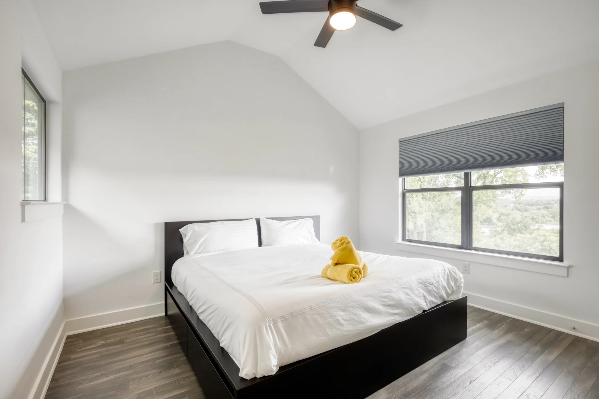 A minimalist bedroom with a white bed, black frame, two pillows, and yellow towels on top. The room has a large window with a dark blue blind, white walls, a ceiling fan, and wooden flooring.