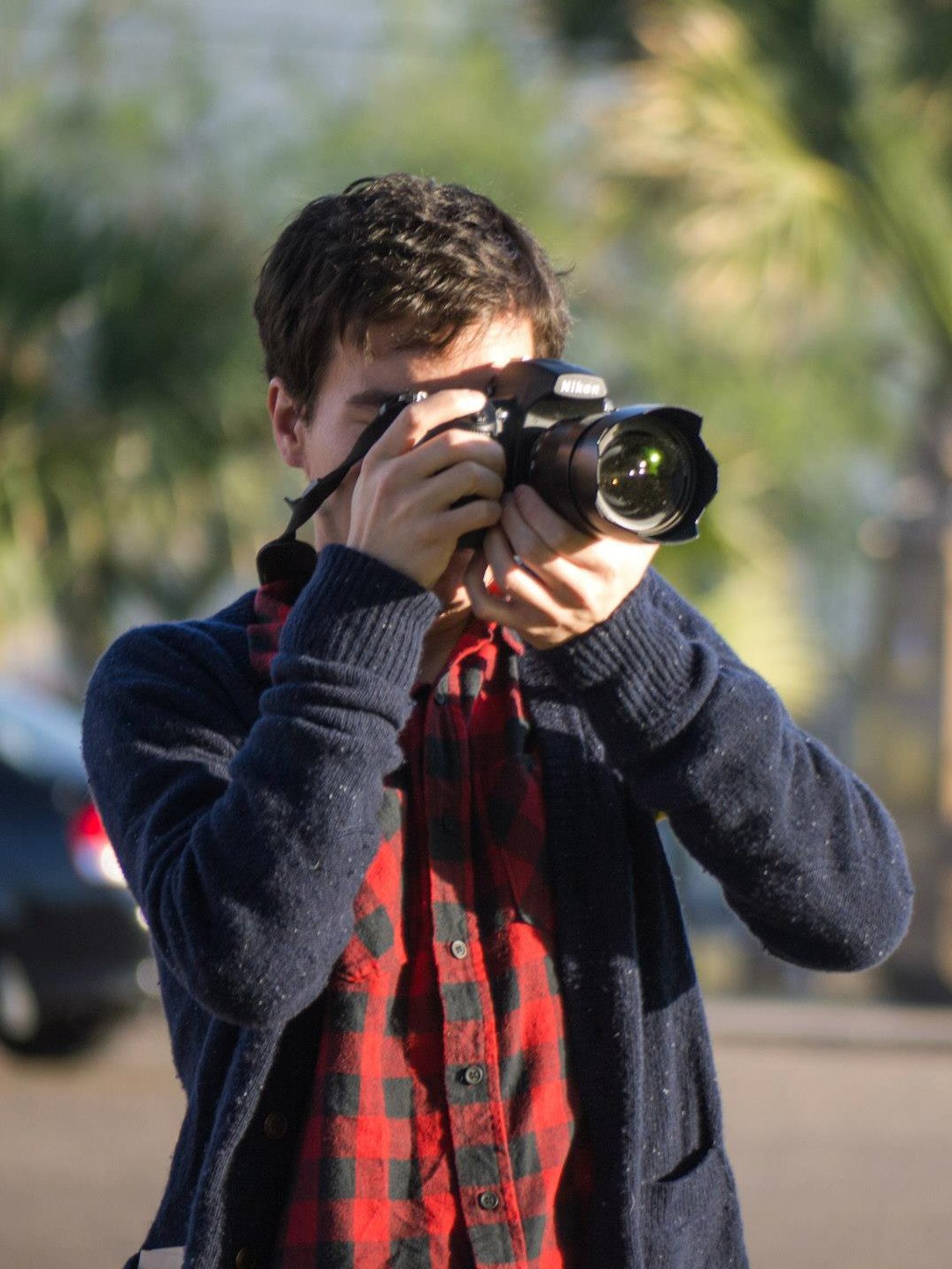 Young man with short brown hair taking a photograph with a Nikon camera outdoors. He is wearing a navy blue sweater over a red and black plaid shirt, with blurred trees and parked cars in the background.