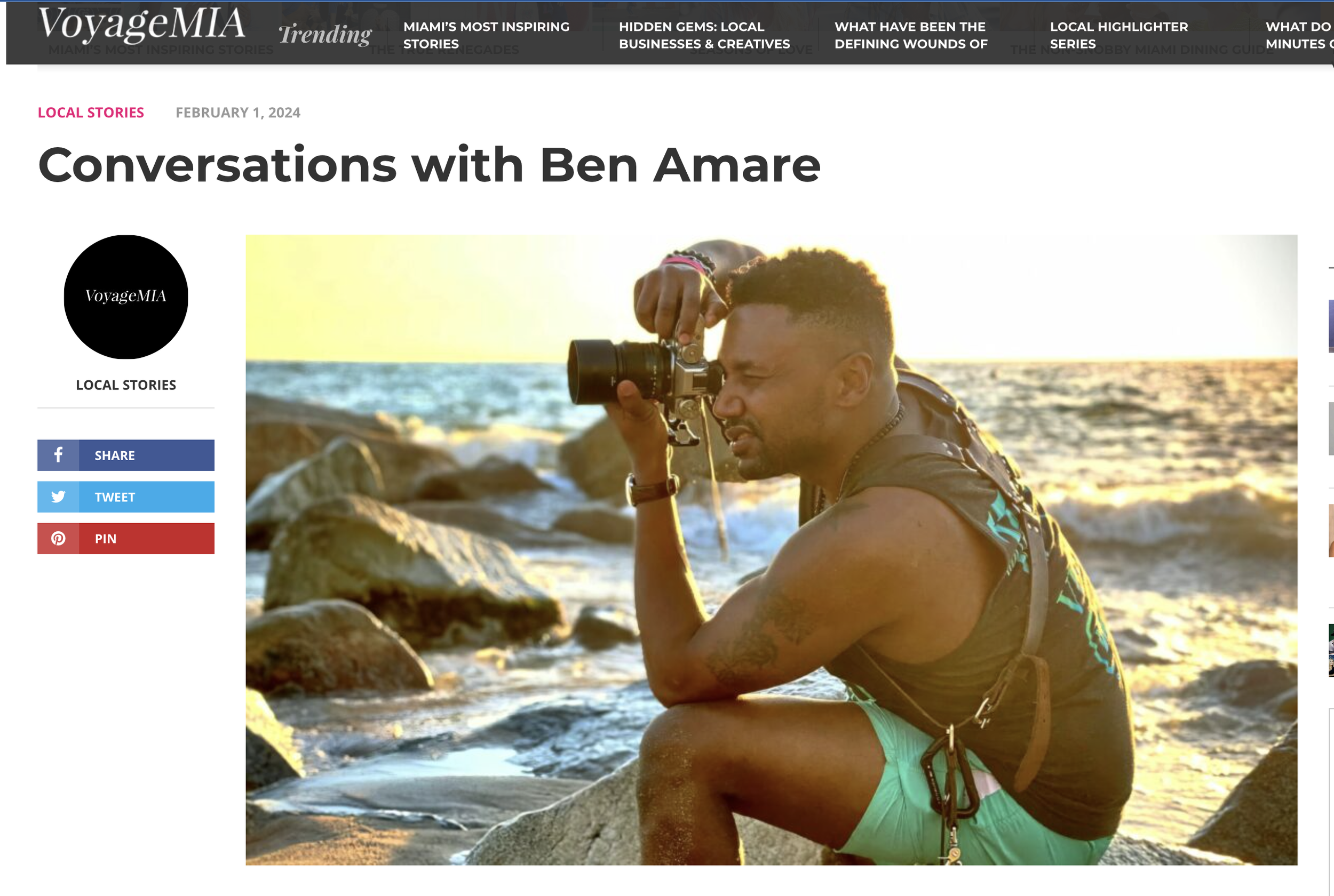 A man with short curly hair and tattoos on his arm sitting on rocks by the ocean at sunset, holding a camera up to his eye, taking a photo.