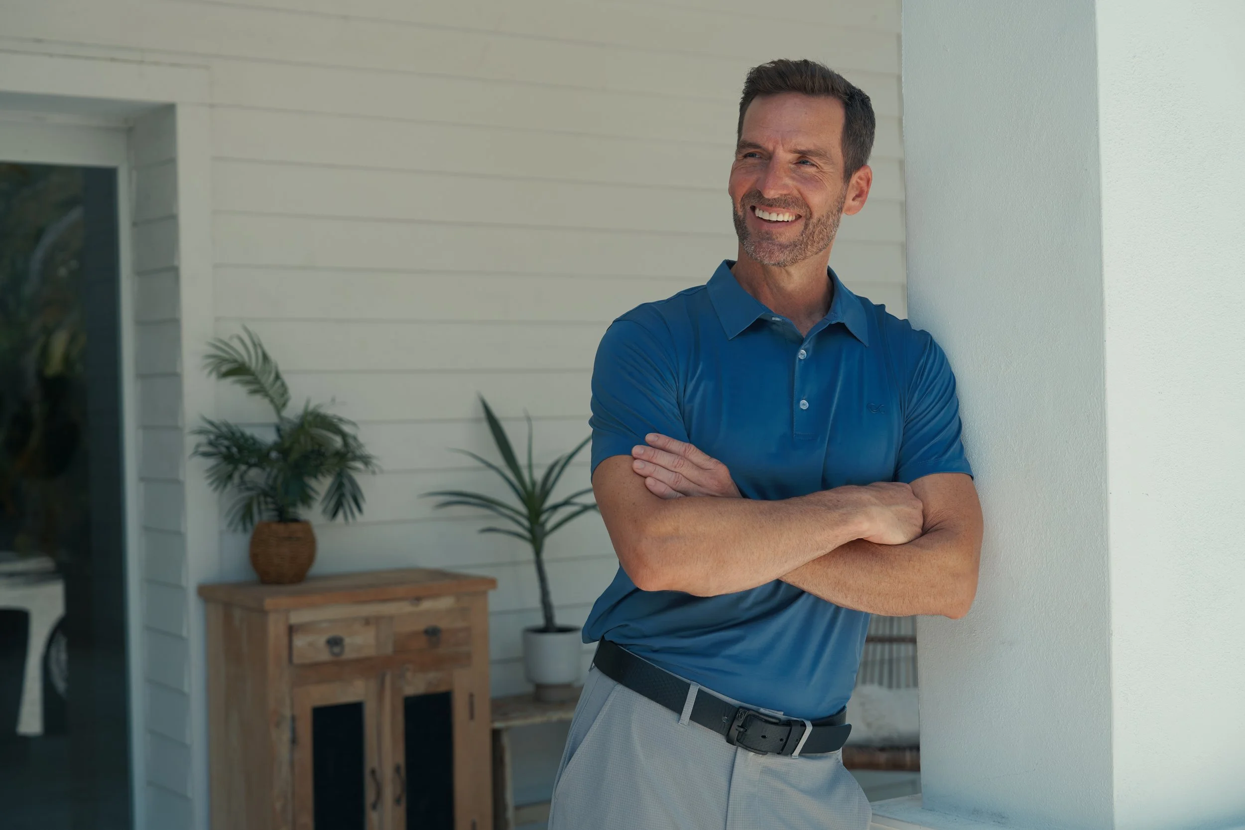 A smiling middle-aged man in a blue polo shirt and light-colored pants standing outdoors, leaning against a white wall with arms crossed.