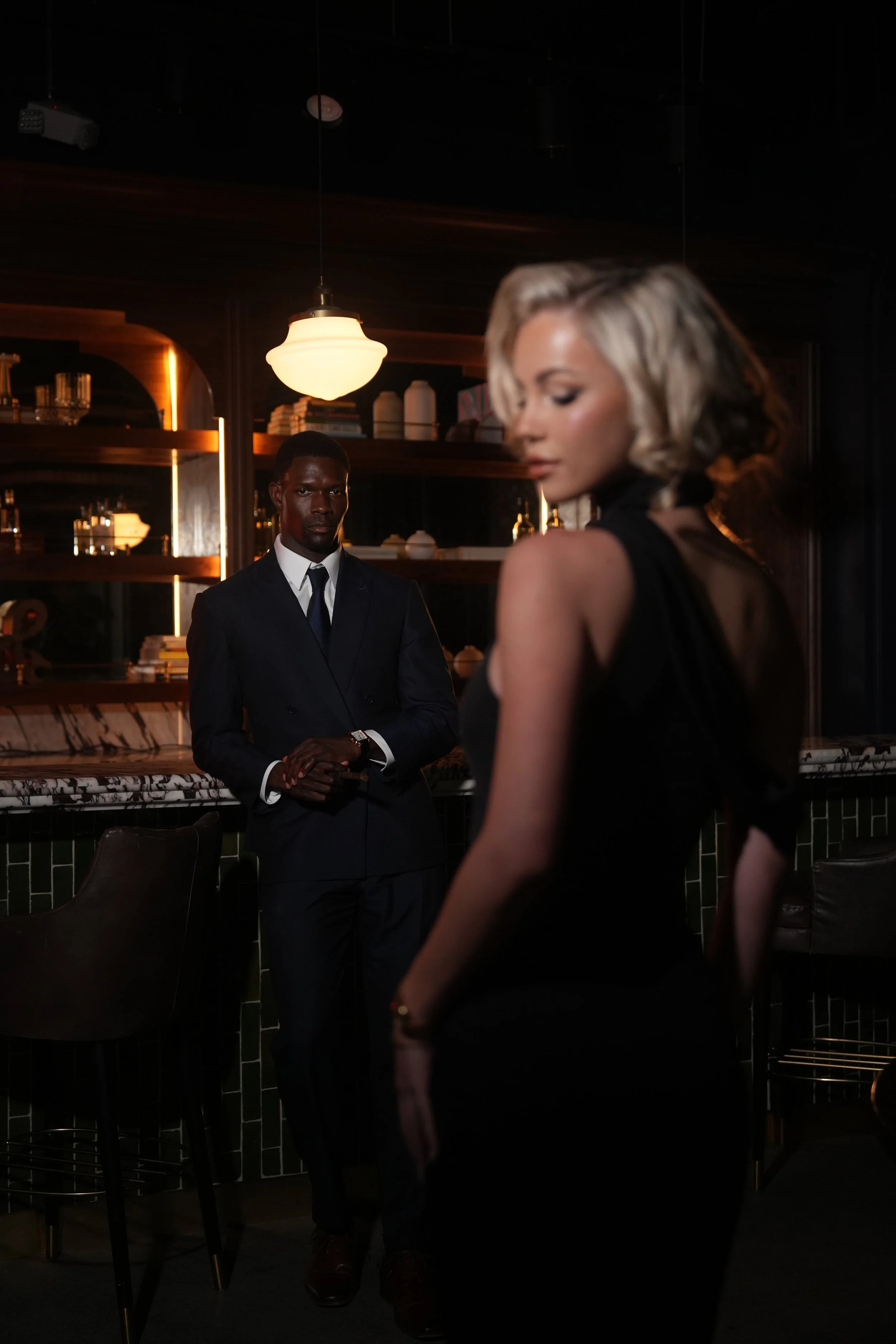 A man in a suit and tie looking at a woman in a black dress in a dimly lit bar or lounge.