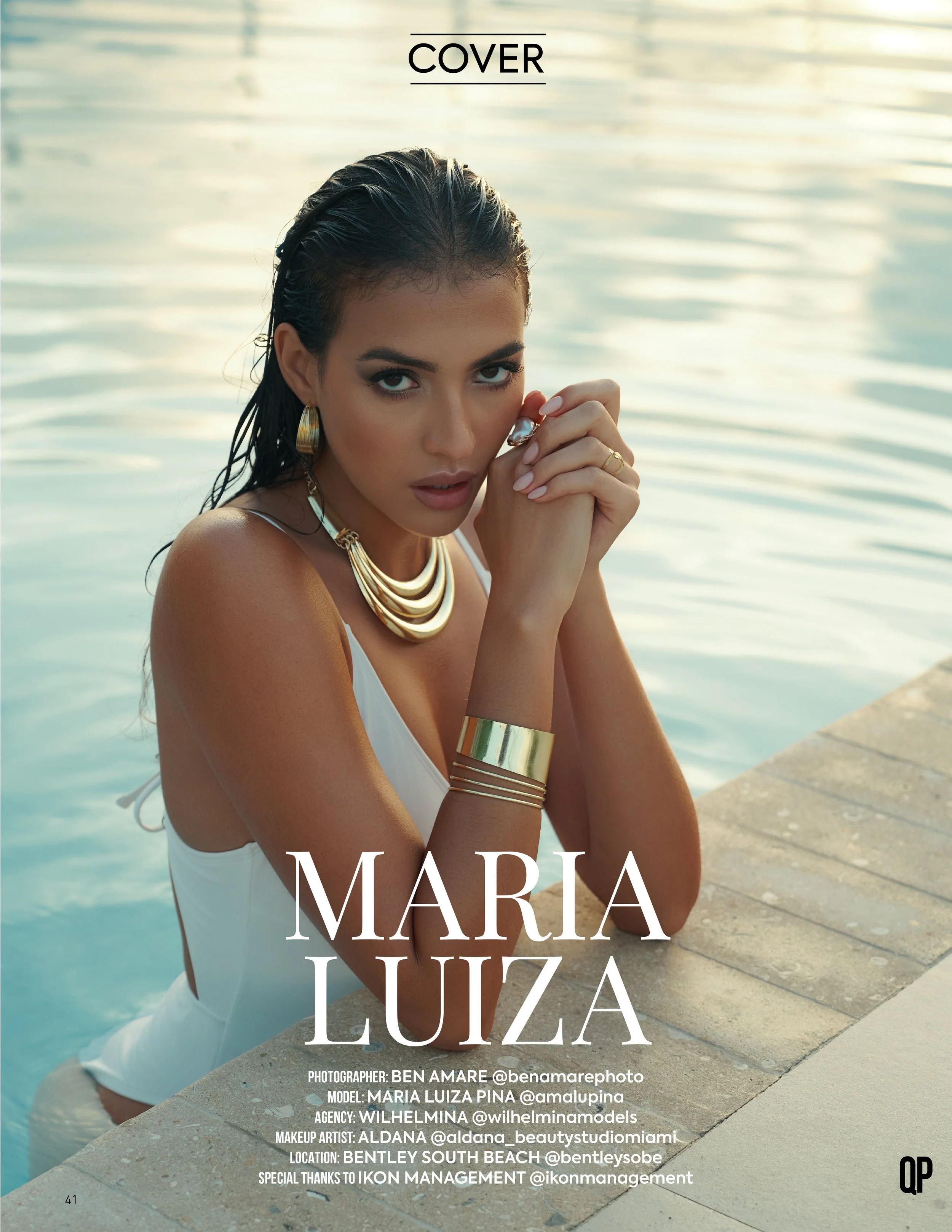 A woman with dark hair and tan skin poses by a pool, wearing gold jewelry and a white swimsuit, looking into the camera with her hands clasped near her face.