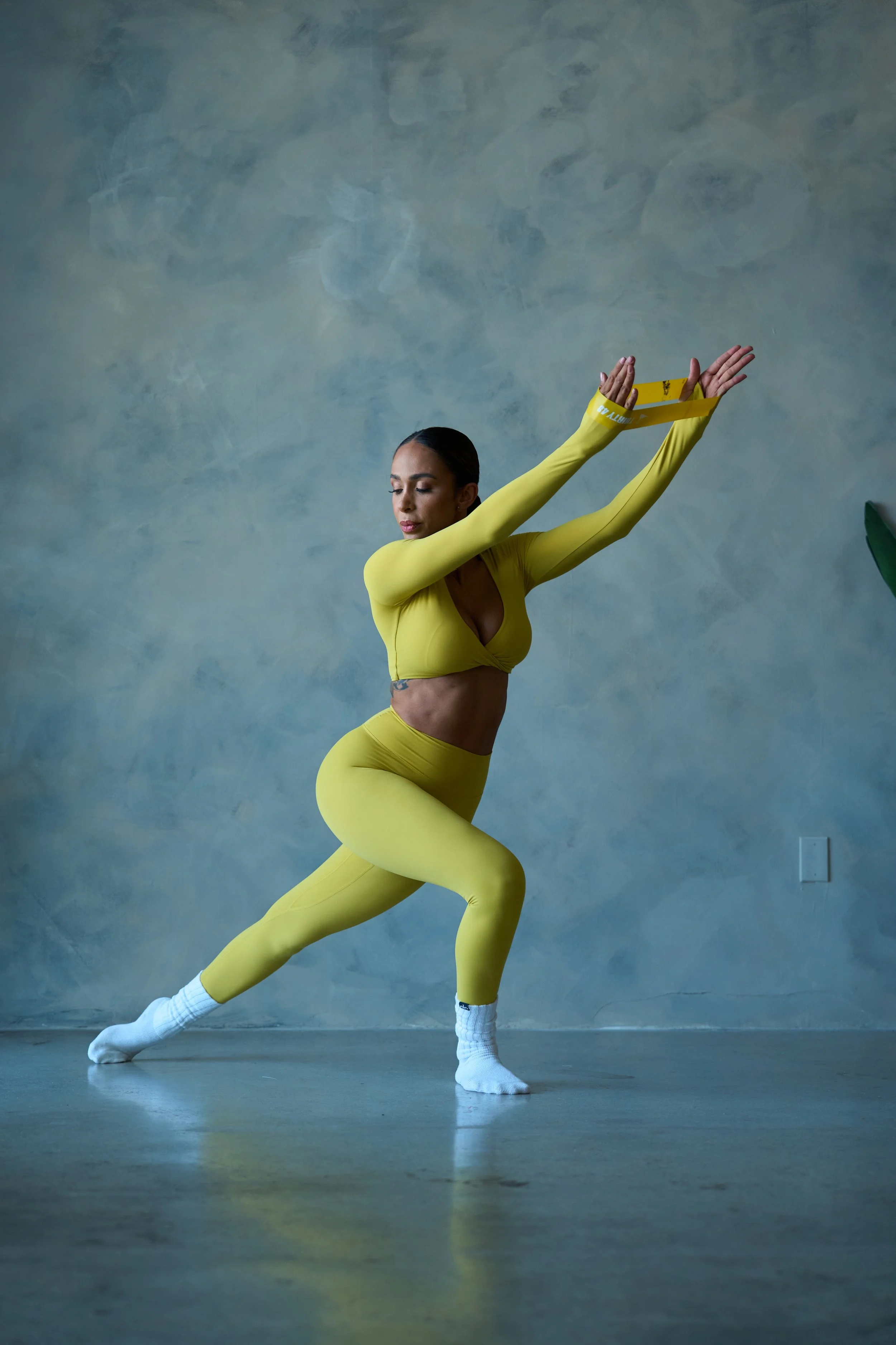 A woman in a yellow athletic outfit stretching indoors against a plain wall.