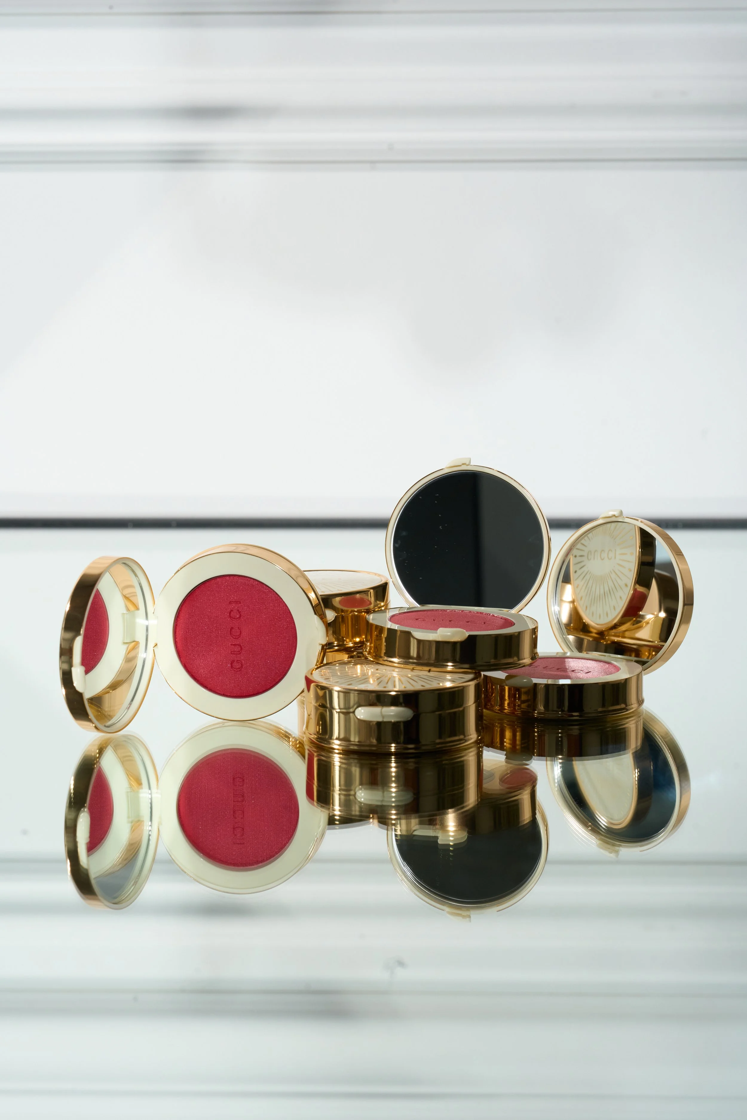 Collection of gold-colored makeup compacts with rose-colored blush or eyeshadow, some open showing product, reflected on a glass surface.