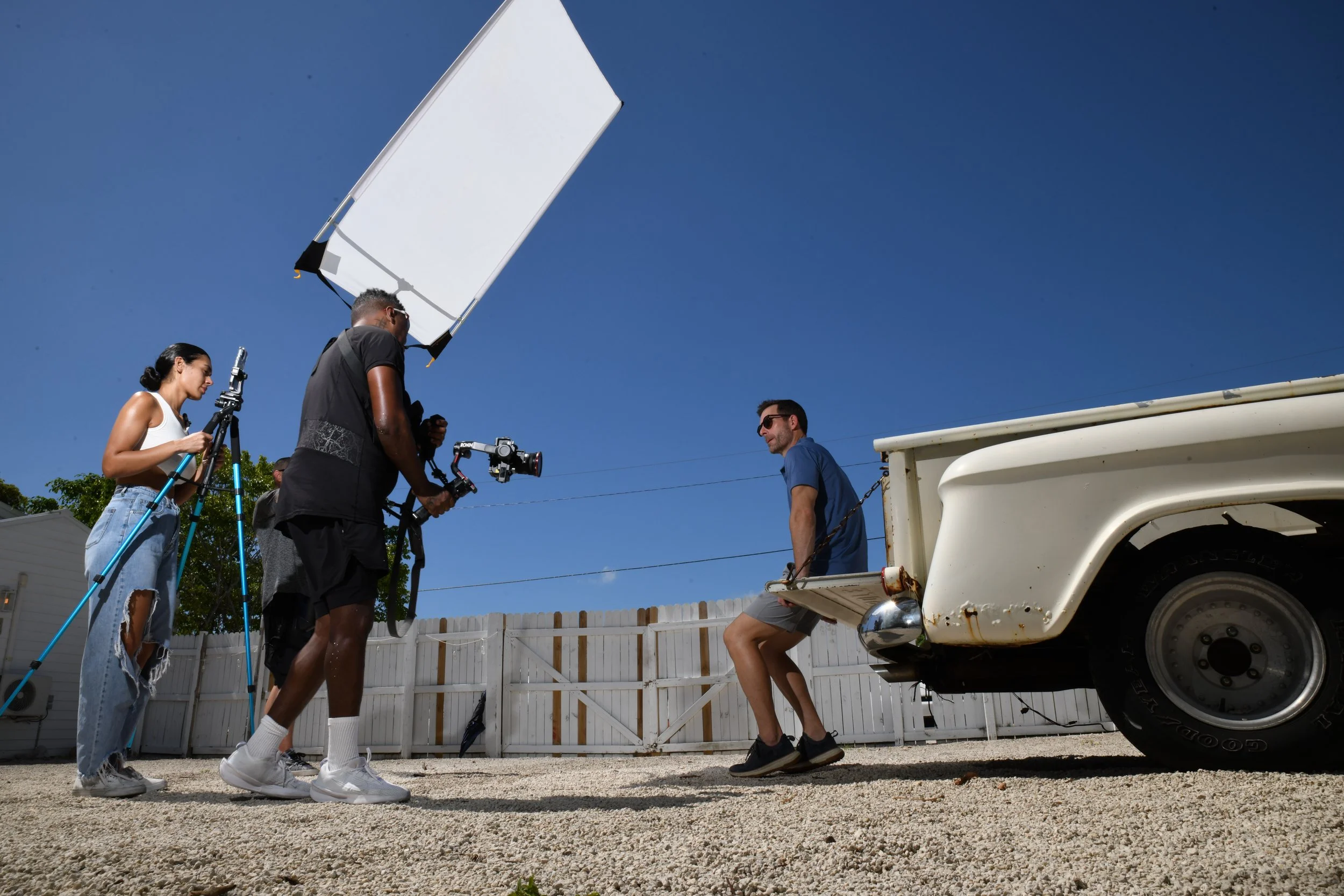 Filming crew recording a man sitting on the back of a vintage truck in a backyard under clear blue sky.