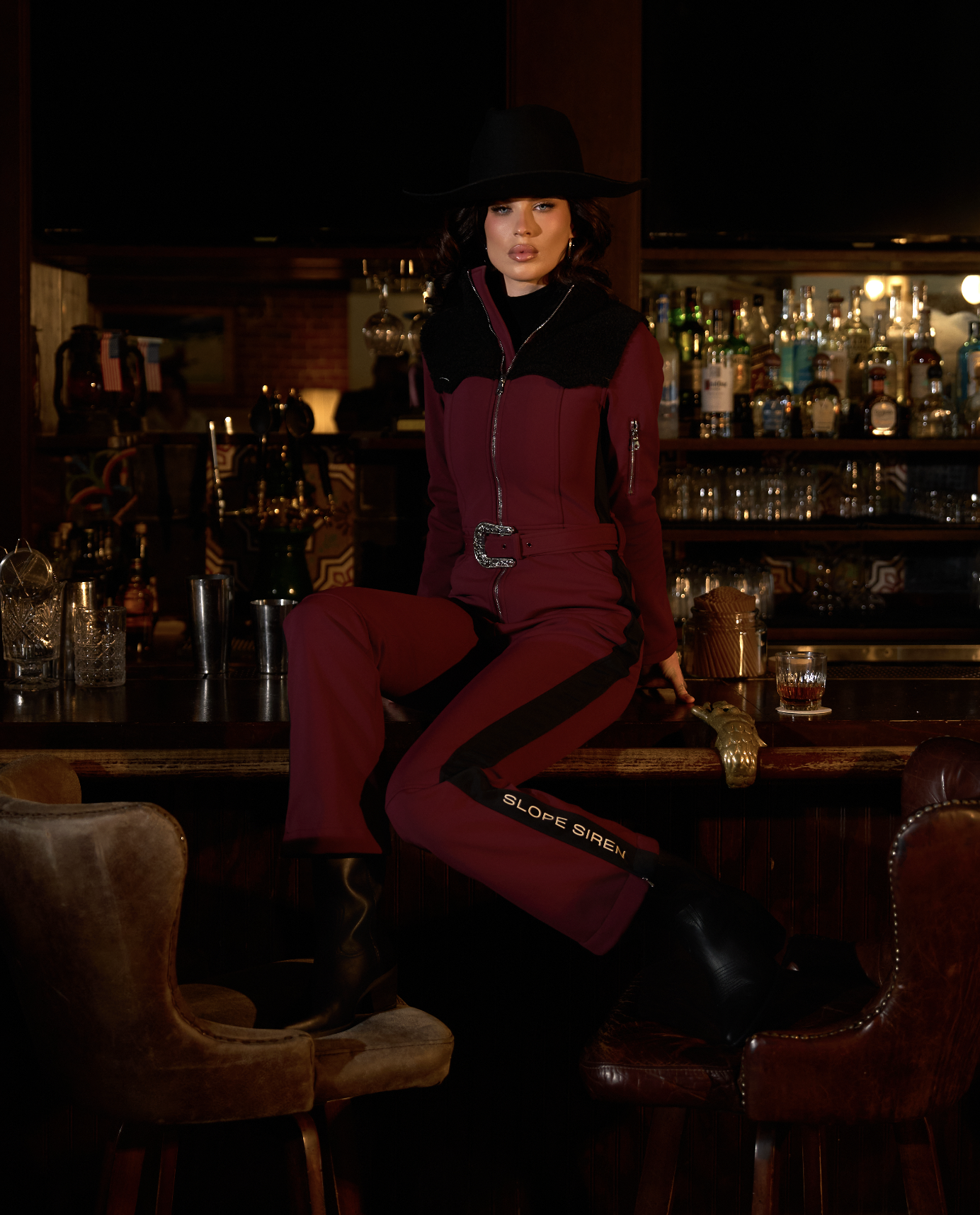 A woman wearing a wide-brimmed black hat and a maroon three-quarter zip jumpsuit with black accents, sitting on a bar counter in a dimly lit bar or lounge.